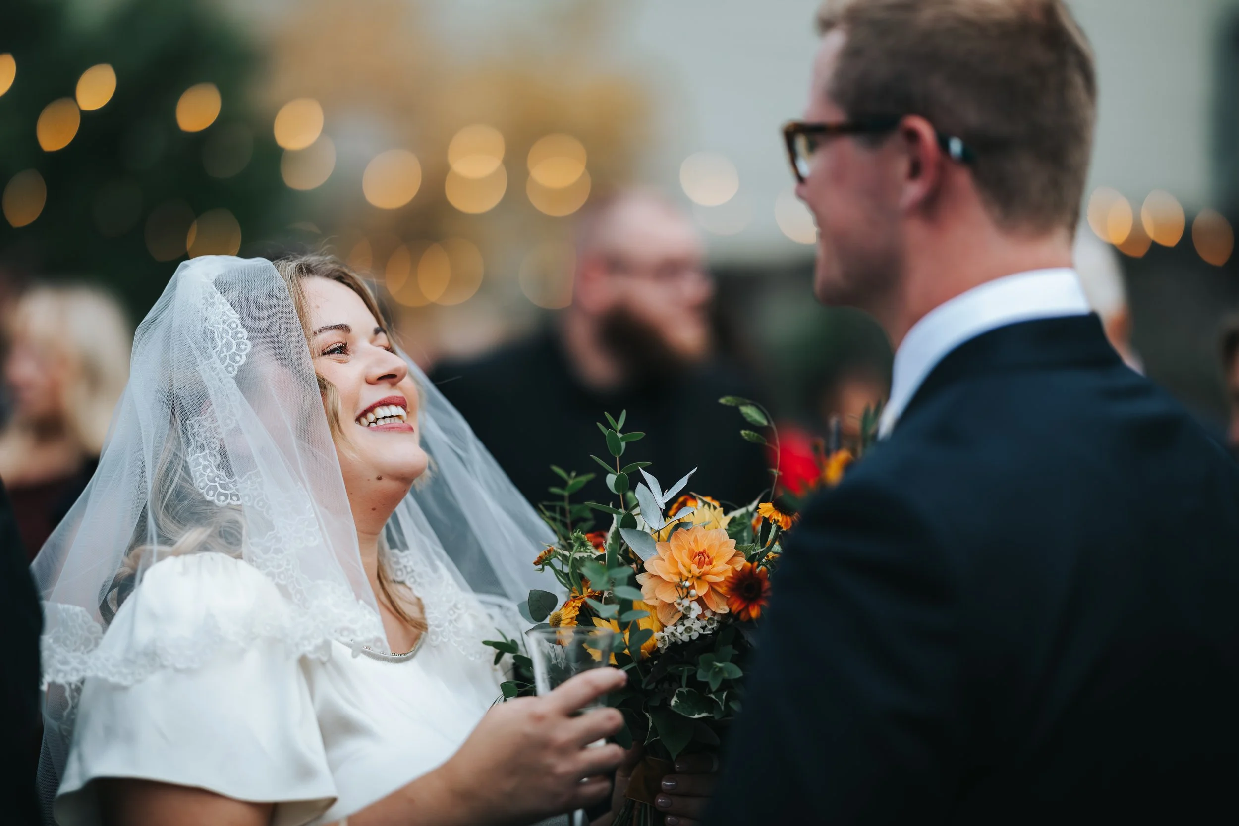 A smiling bride with a lace veil and white dress holding a bouquet of yellow, orange, and white flowers, looking at a groom in a suit and glasses during a wedding celebration