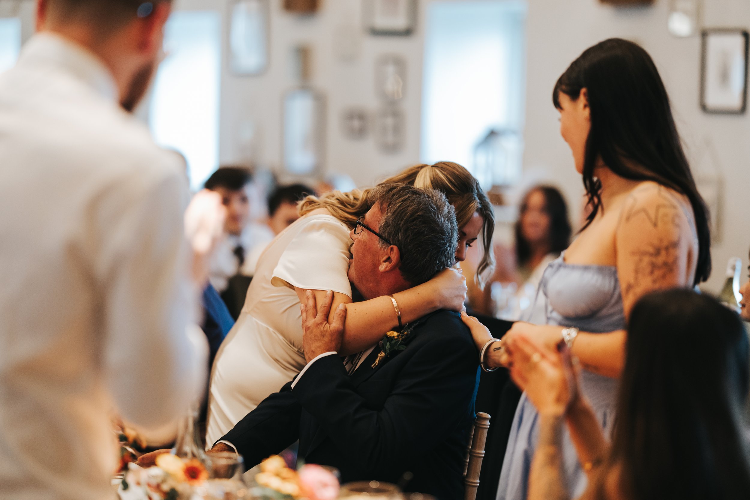 A woman is hugging and kissing an older man at a formal event, possibly a wedding, with other guests in the background. The woman has blonde hair and is wearing a white dress. The older man is seated, wearing a dark suit and glasses, and is being emb