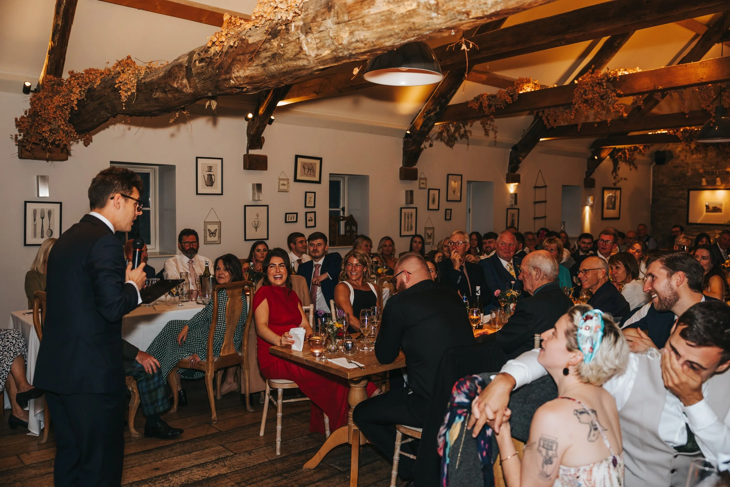 A man in a black suit speaking into a microphone at a wedding reception, with guests seated at tables laughing and listening in a decorated rustic venue.