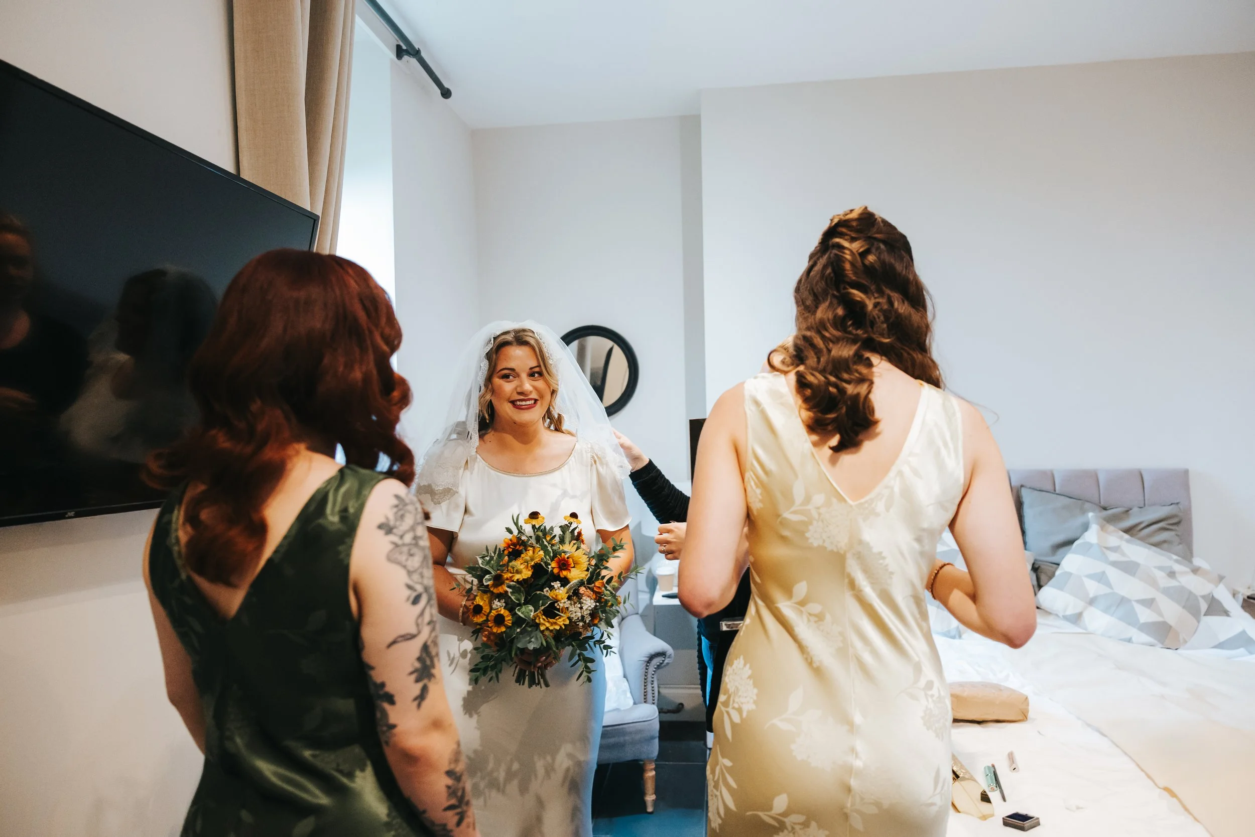 A bride holding a bouquet of flowers, smiling, with three women around her, in a bedroom.