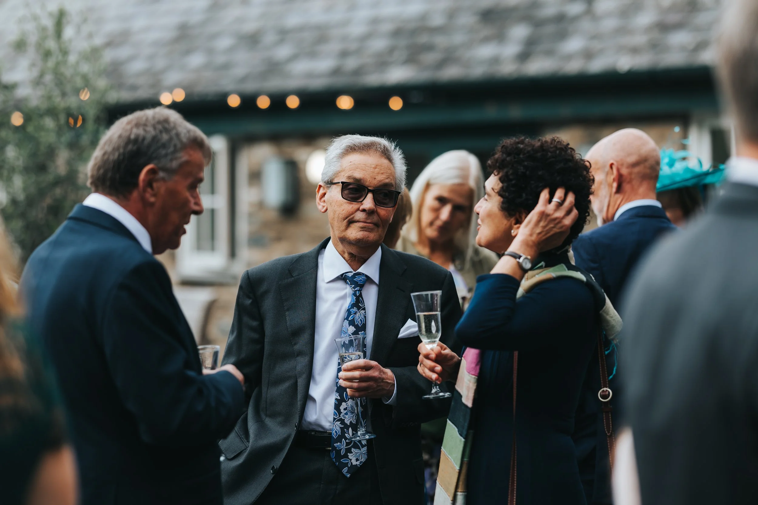 Group of people at an outdoor gathering, dressed in formal attire, holding champagne glasses, with a stone building in the background.