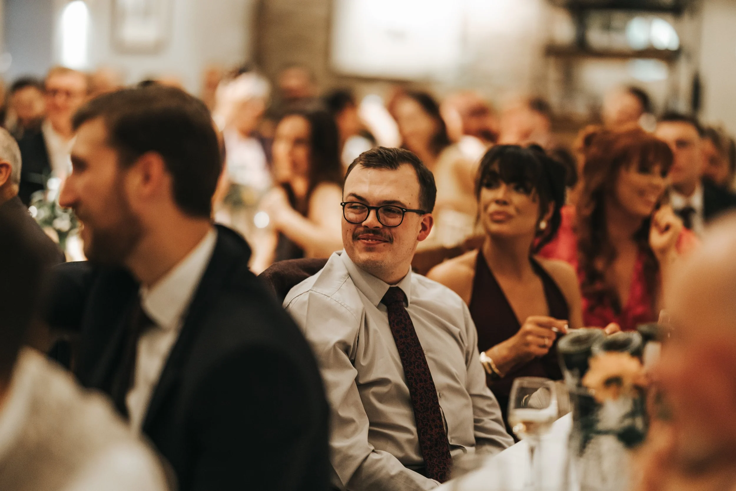 A man with dark hair, glasses, and a mustache is smiling and looking to his right during a formal event, with a crowd of people in the background.