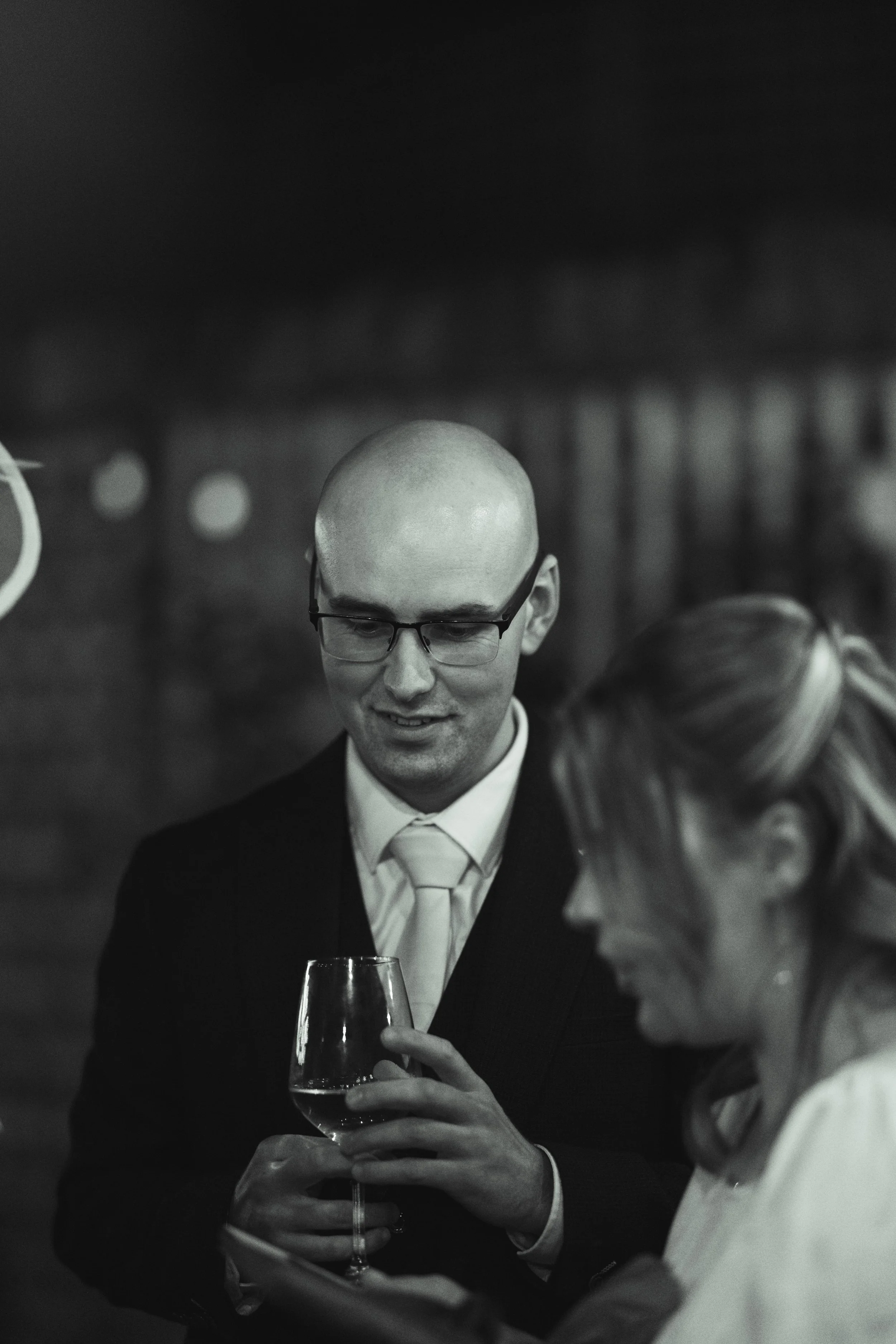 A man in a suit and glasses holding a wine glass and smiling while talking to a woman at a social event in black and white.