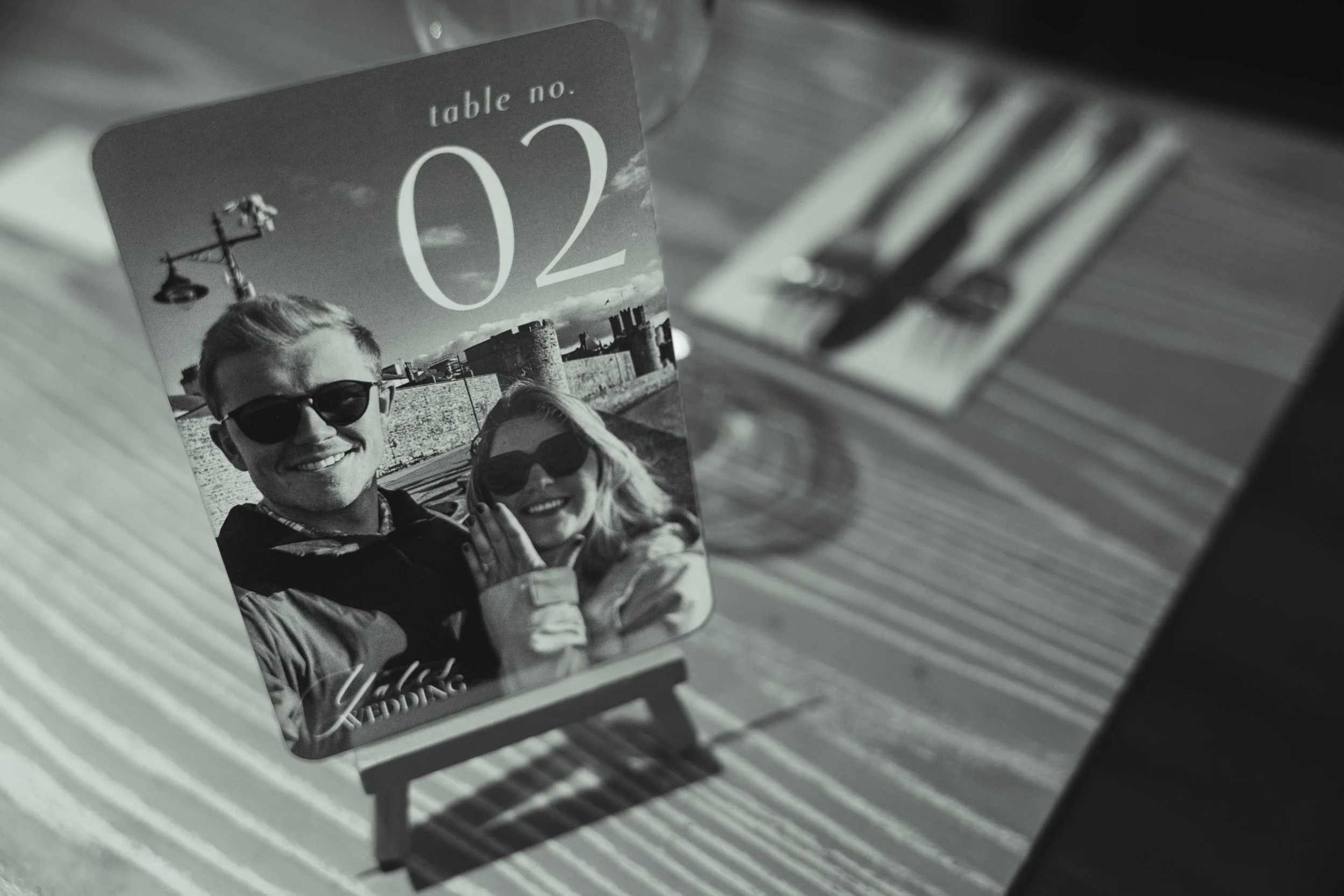 A table number sign for a wedding, with a black and white photograph of a smiling man and woman wearing sunglasses, against an outdoor castle backdrop. The sign displays the number '02' and the word 'WEDDING' at the bottom.