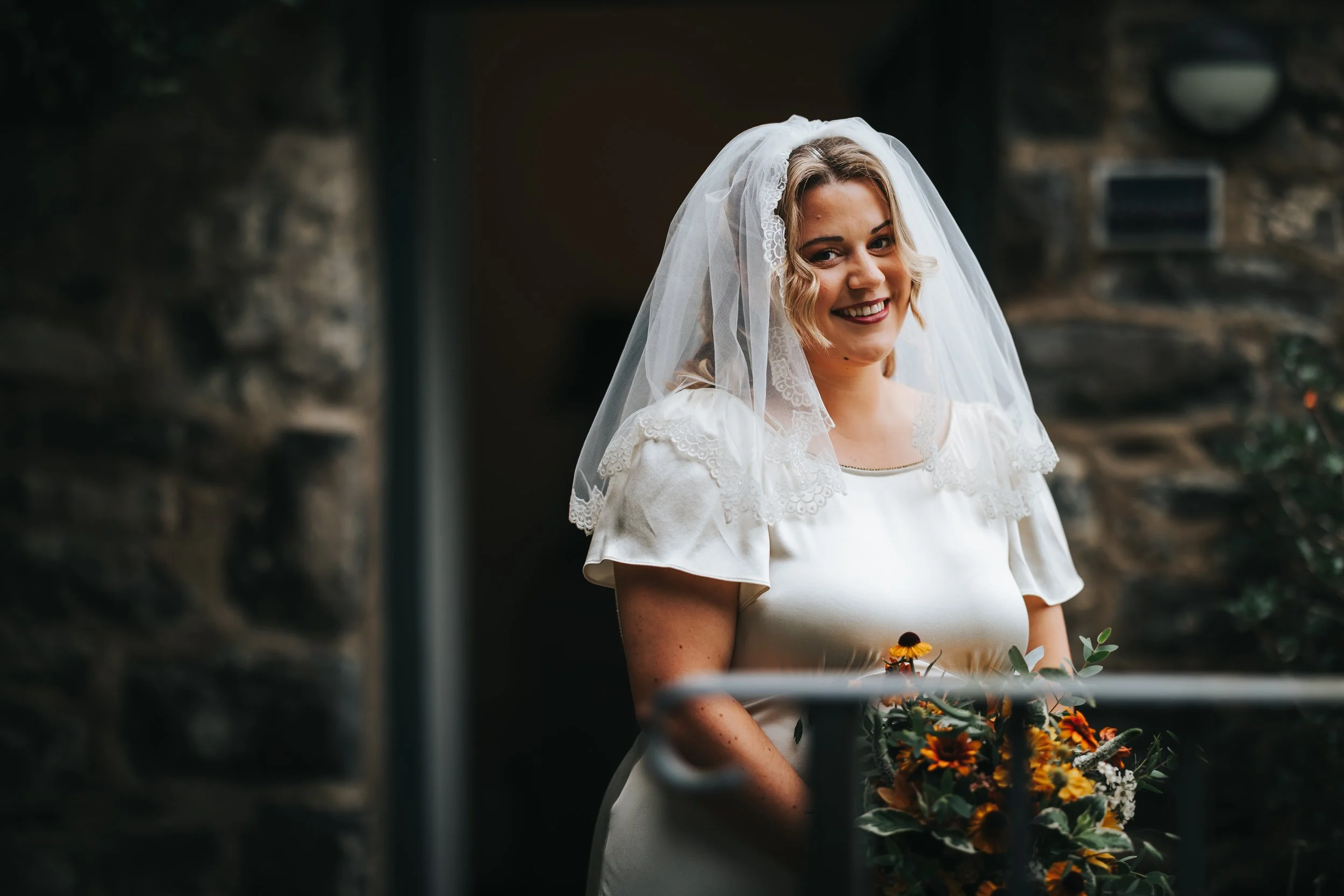 A smiling woman in a vintage wedding dress with lace details and a veil, holding a bouquet of yellow, orange, and white flowers, standing in front of a stone wall.