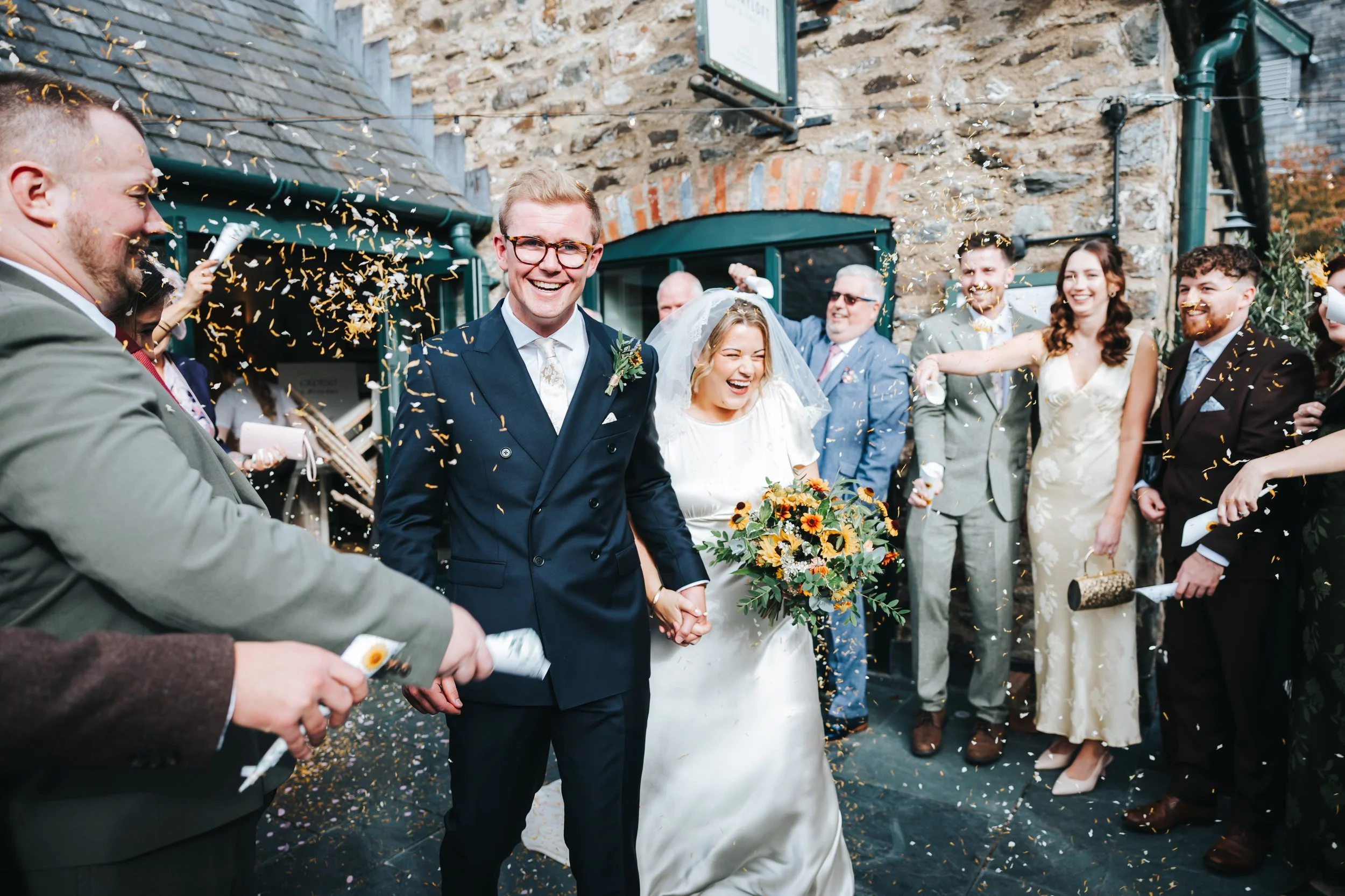 Happy bride and groom holding hands in the center, surrounded by friends and family celebrating with confetti and laughter outside a rustic stone building.