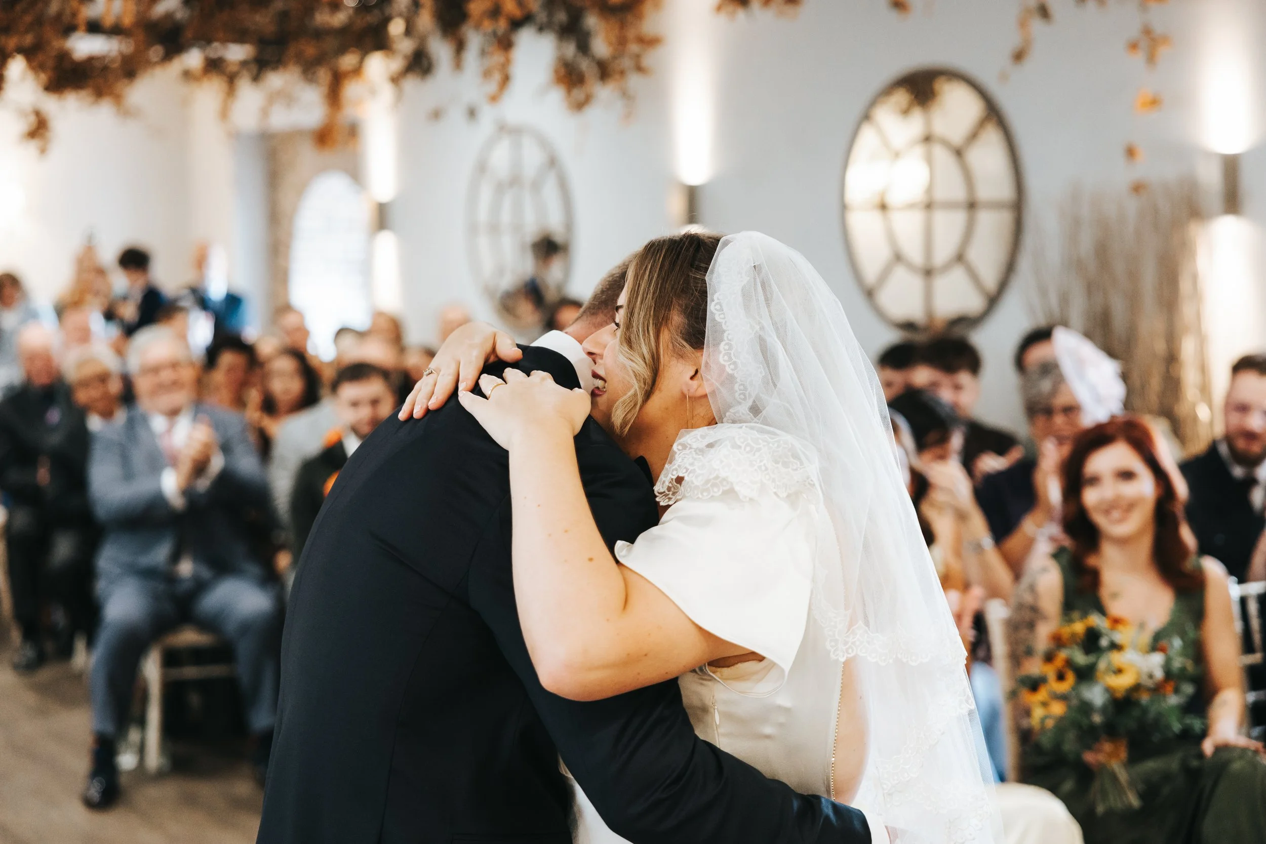 A bride and groom embrace and share a kiss during their wedding ceremony, with guests clapping and smiling in the background.