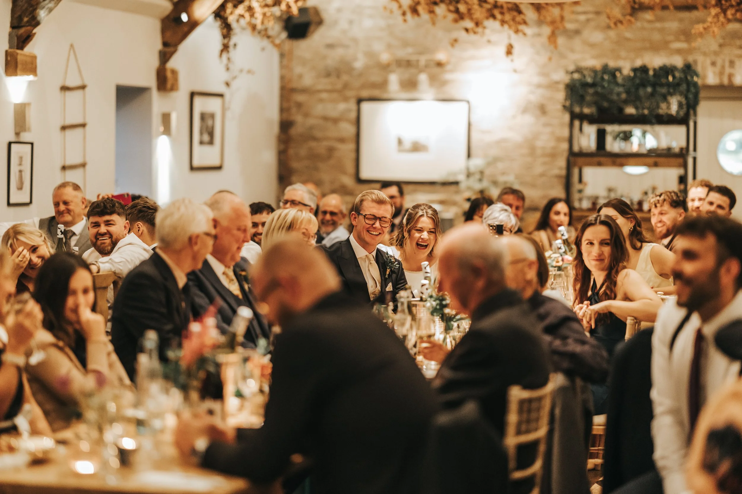 A group of wedding guests sitting at long tables, smiling and laughing during a celebration in a warmly lit rustic venue with wooden beams and brick walls.