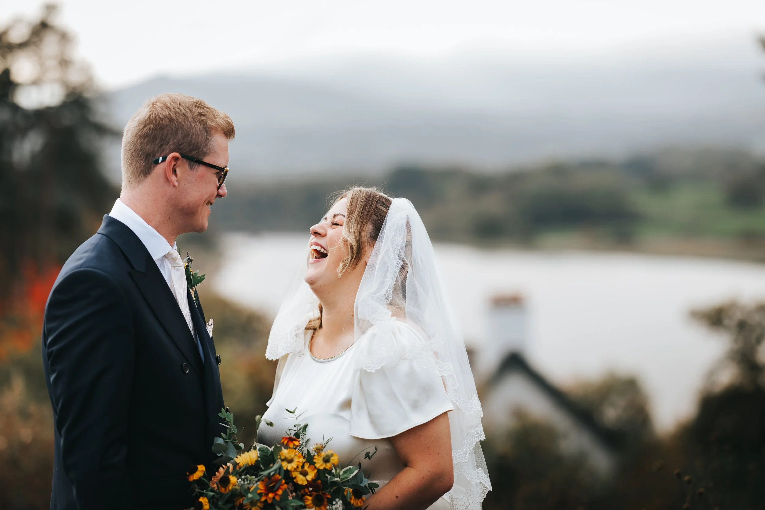 A bride and groom at their wedding ceremony outdoors, smiling and laughing together, with a lake and mountains in the background.