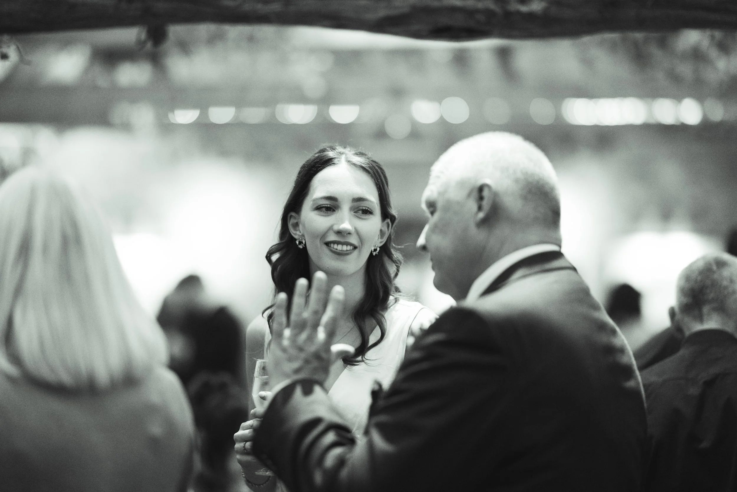 A woman with dark wavy hair smiling and talking to a man with short gray hair at a formal event. The woman is holding a glass of wine, and there are other people in the background.