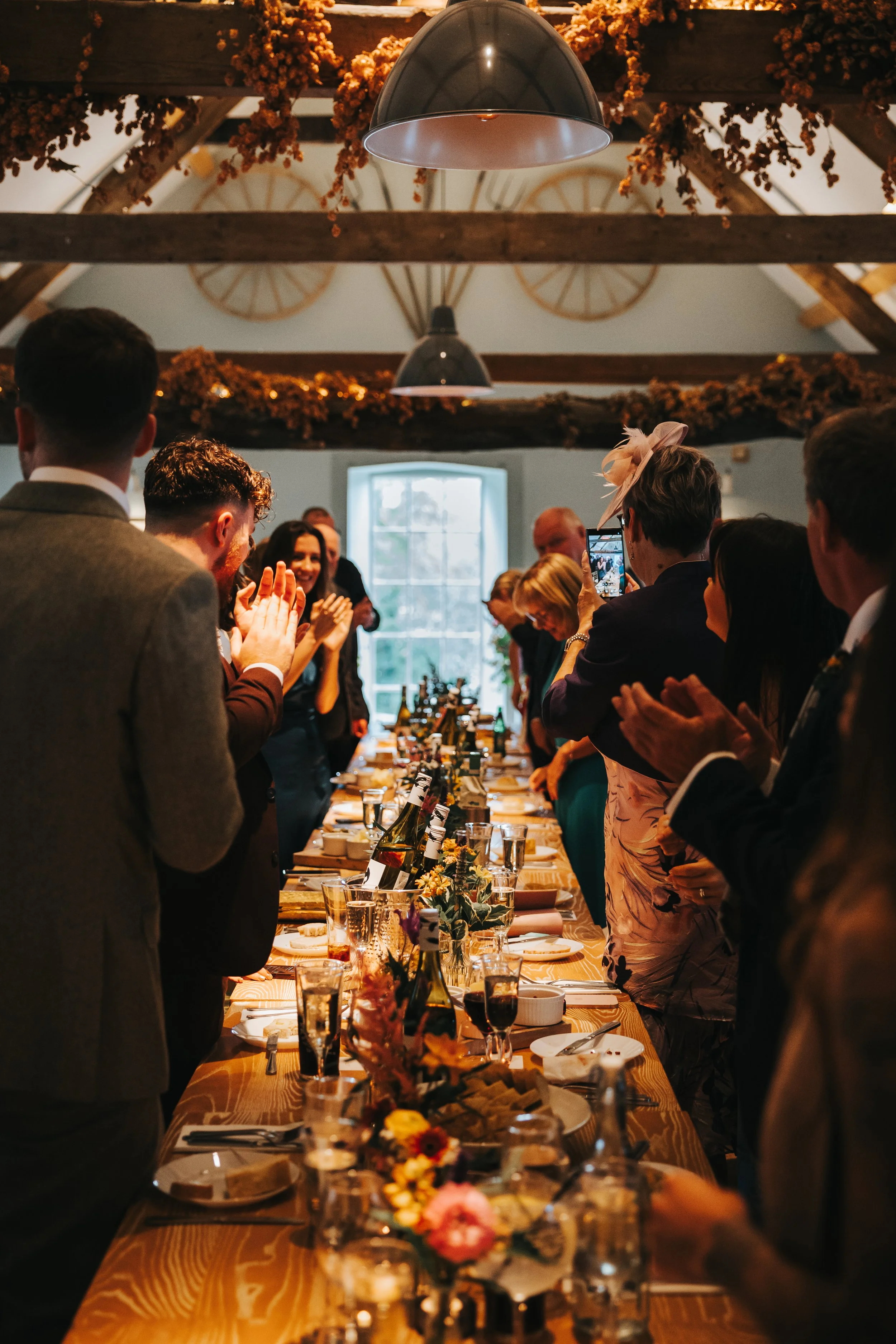 People gathered around a long decorated dining table at a celebration, in a rustic venue with wooden beams, flowers, and a window in the background.
