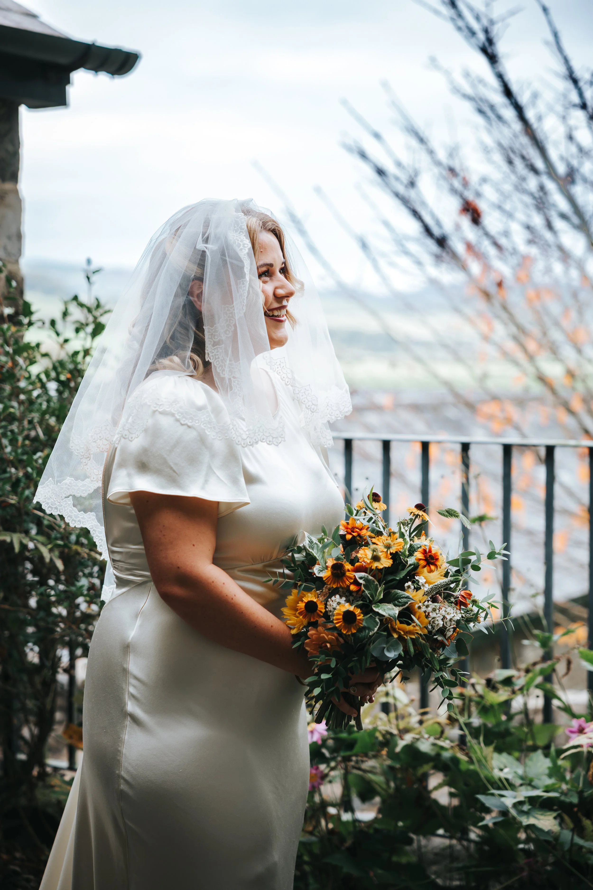 A smiling bride in a white satin dress and lace veil holding a bouquet of sunflowers and greenery, standing outdoors near a balcony with trees and a scenic landscape in the background.