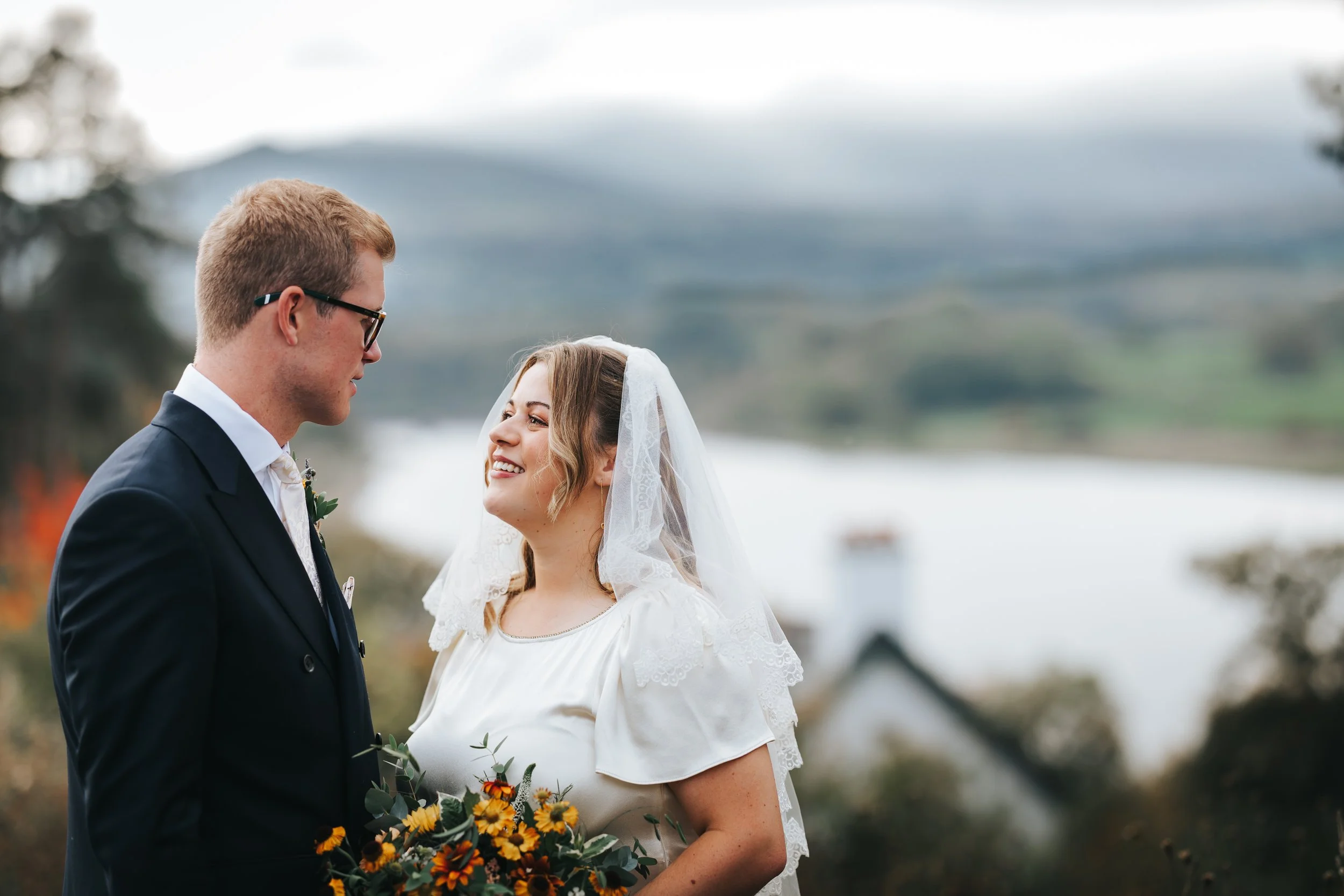 A bride and groom standing outdoors with a lake and mountains in the background, smiling at each other.