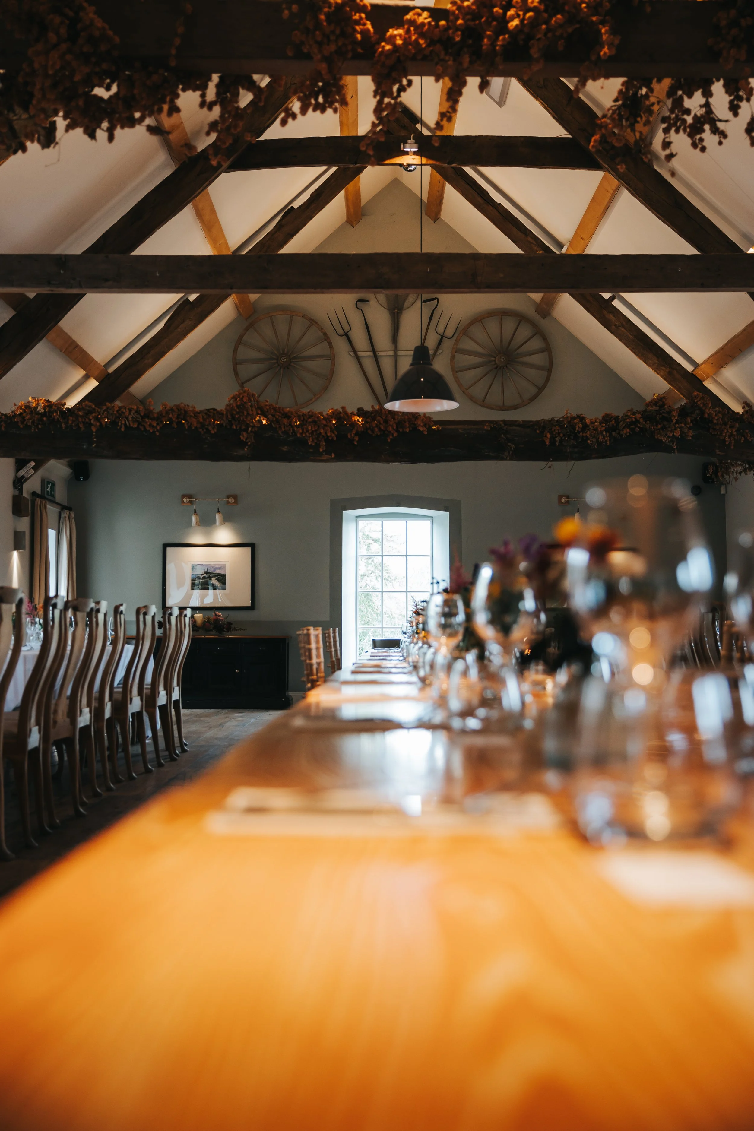 A rustic dining room with a long wooden table set for a meal, with many chairs around it. The room has a high vaulted ceiling with exposed wood beams, decorated with hanging flowers. Two large wagon wheels and farming tools are mounted on the wall ne