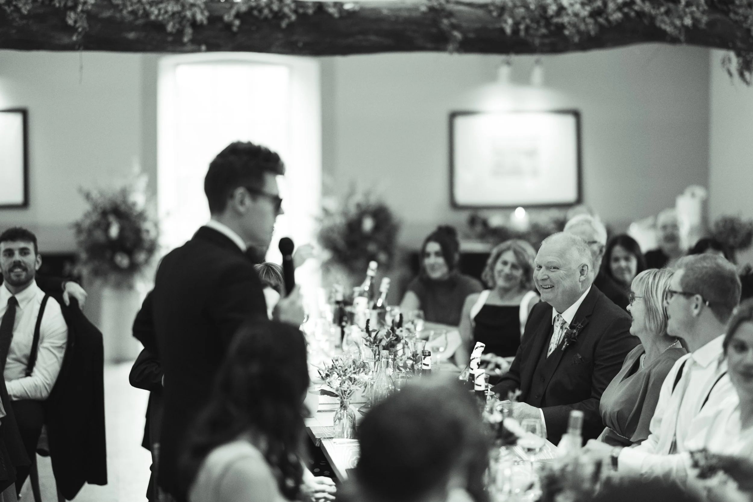 A black and white photo of a man in a suit giving a speech at a formal gathering, with people seated at a table smiling and listening.