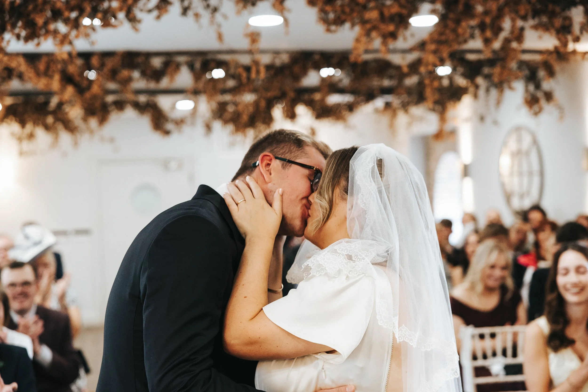 A couple, dressed in wedding attire, shares a kiss at their wedding ceremony with guests seated in the background.