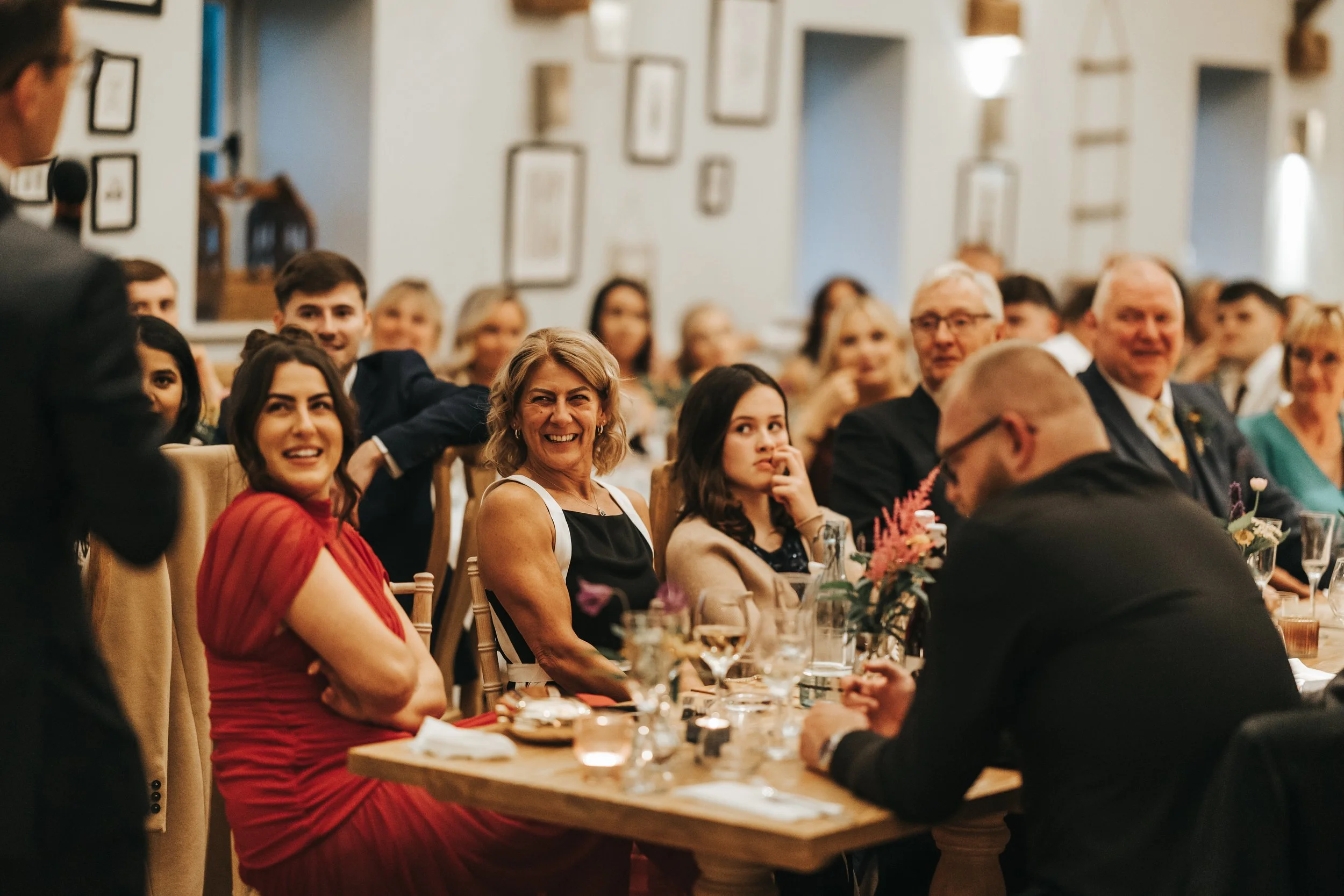 People attending a formal event or celebration, sitting at a table with flowers and drinks, with a speaker in the foreground, smiling and engaging with the speaker.