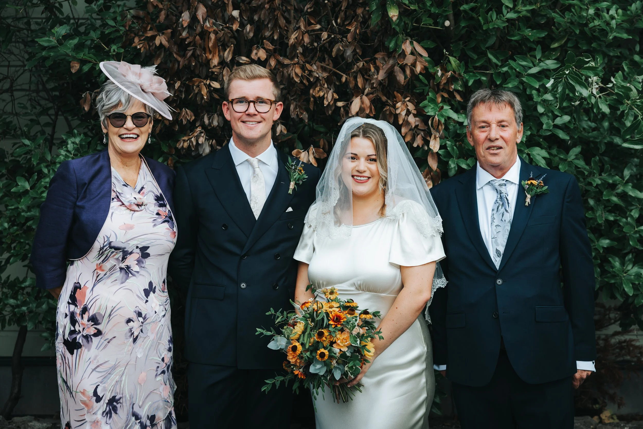 Group of five people dressed in formal wedding attire, standing outdoors in front of a leafy background. The bride, holding a bouquet, is in the center. To her left are a groom in a dark suit and a woman in a floral dress with a hat. To her right are
