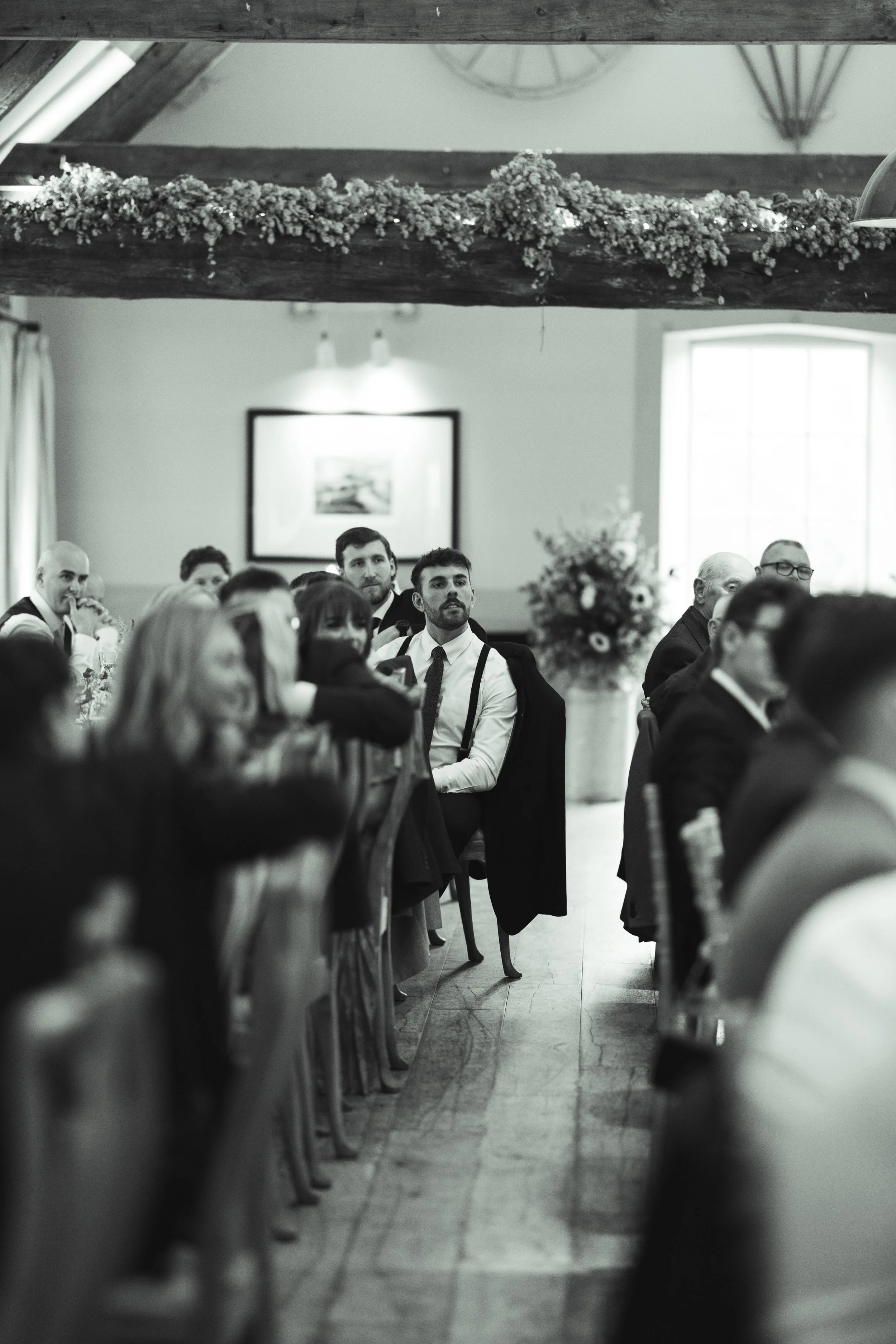 People attending a formal event, seated at long tables in a room with wooden beams and a large window, with a man in a white shirt and suspenders sitting in the middle of the image, looking attentively.