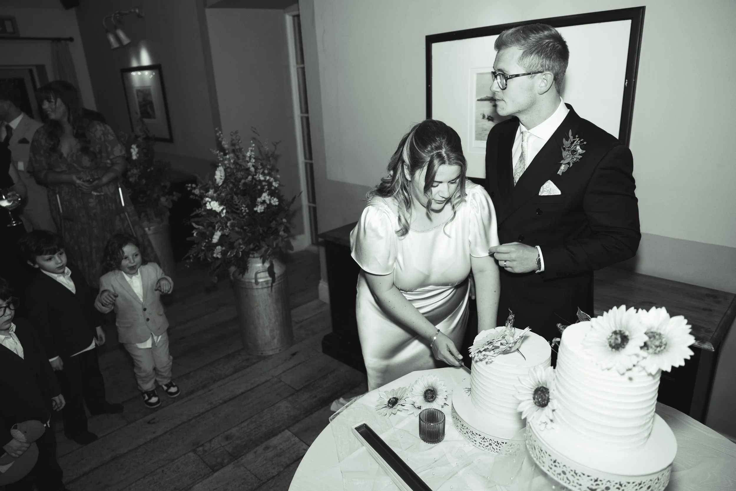 A bride and groom cutting a wedding cake with floral decorations, inside a decorated reception hall with guests watching.
