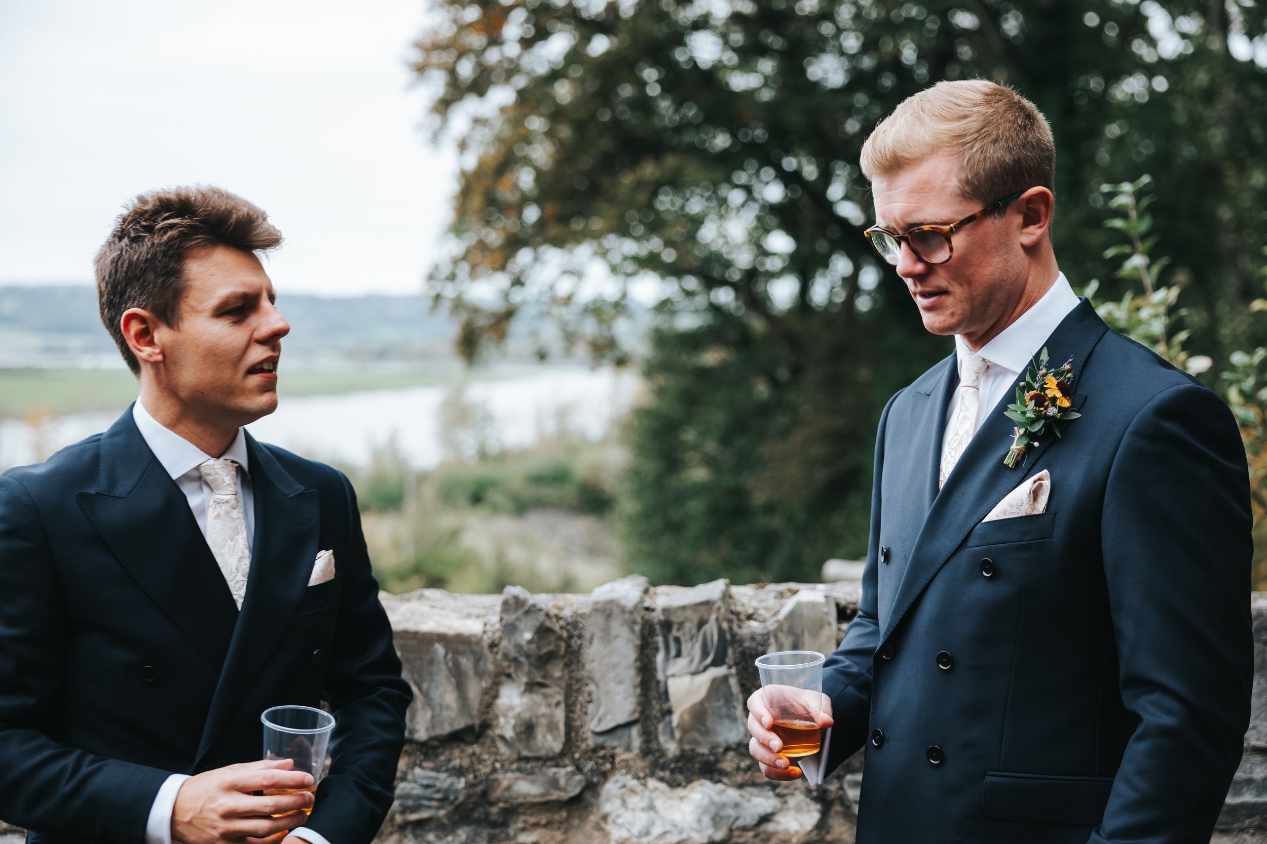 Two men in suits standing outdoors near a stone wall, holding drinks, engaged in conversation during a wedding or formal event.
