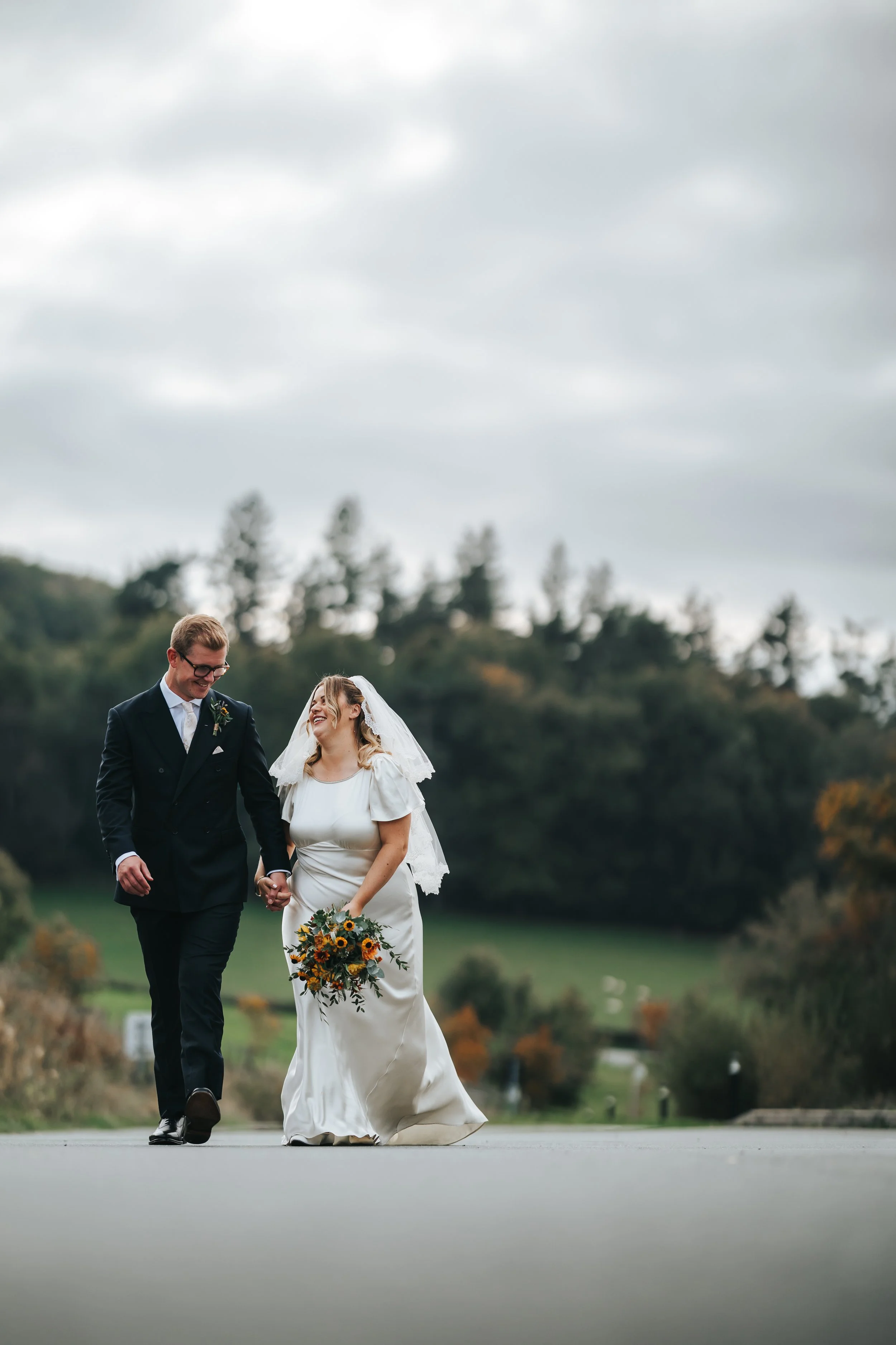 A bride and groom walking outdoors on a cloudy day, holding hands and smiling. The bride is wearing a white wedding dress and veil, carrying a bouquet of flowers. The groom is in a dark suit and glasses. The background shows trees and a slightly hill