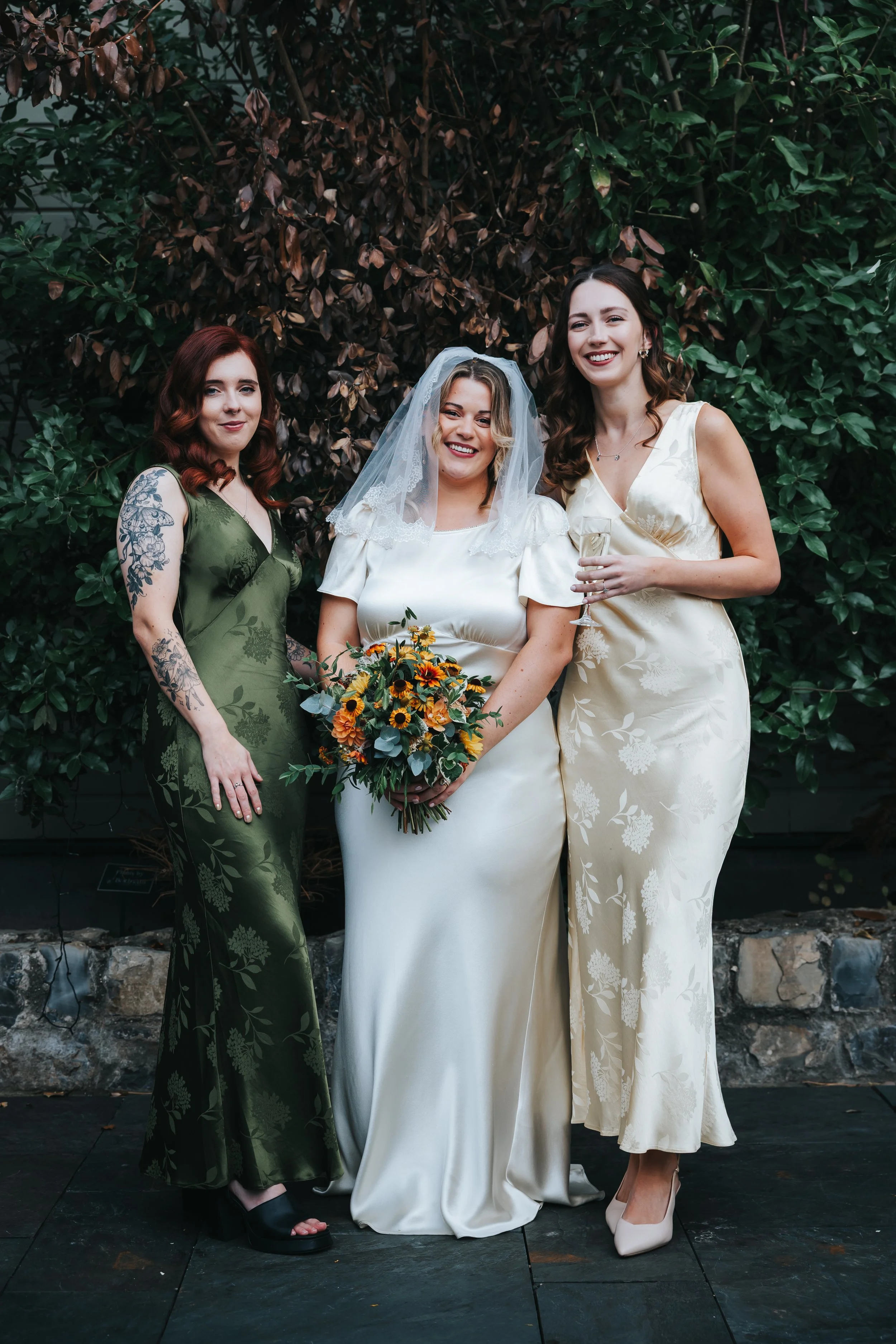 Three women at a wedding, one in a white wedding dress holding a bouquet, and two in elegant dresses, standing outdoors in front of greenery.