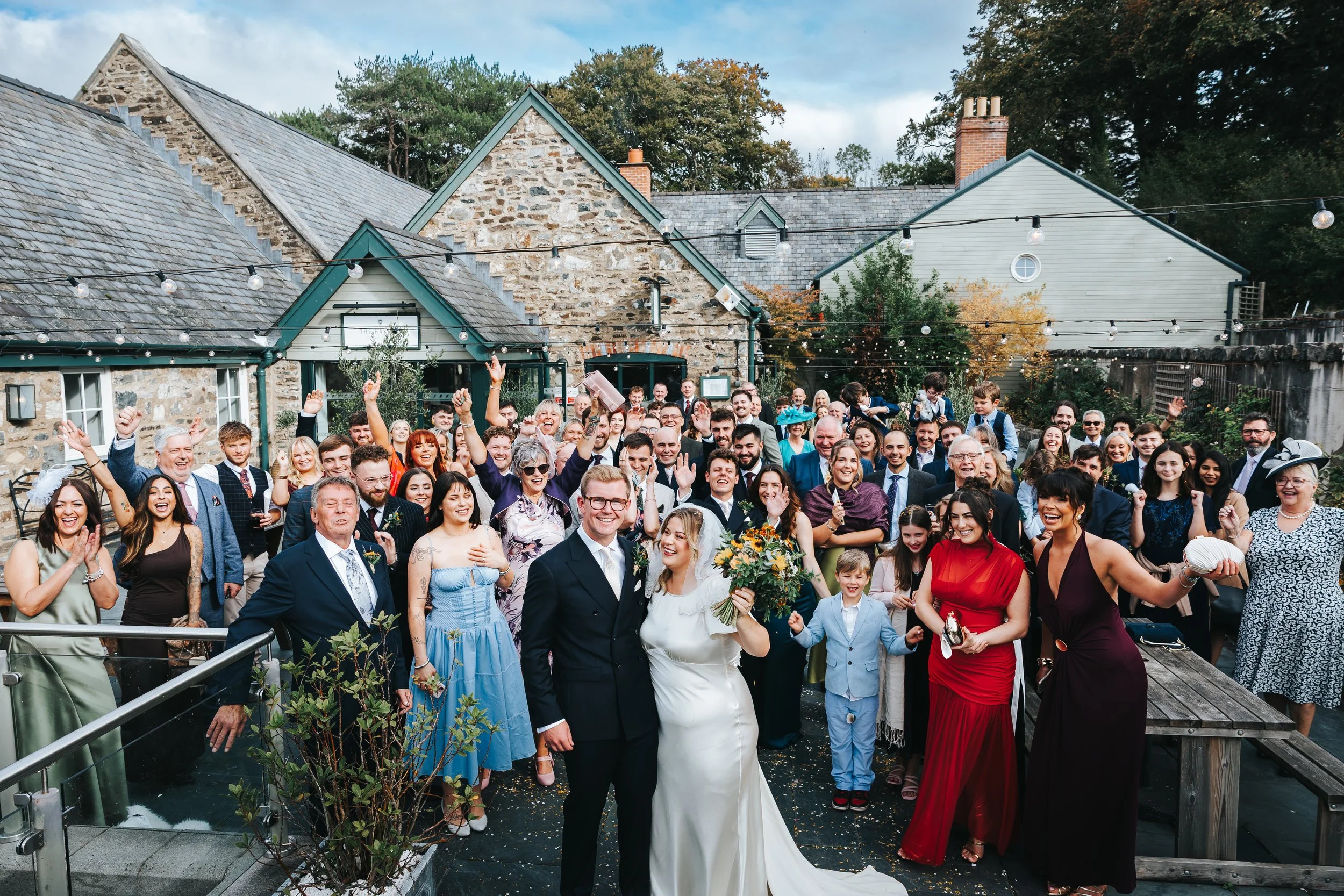 A large wedding celebration outdoors in front of a rustic stone and wood building, with the bride and groom at the center surrounded by guests smiling and celebrating.