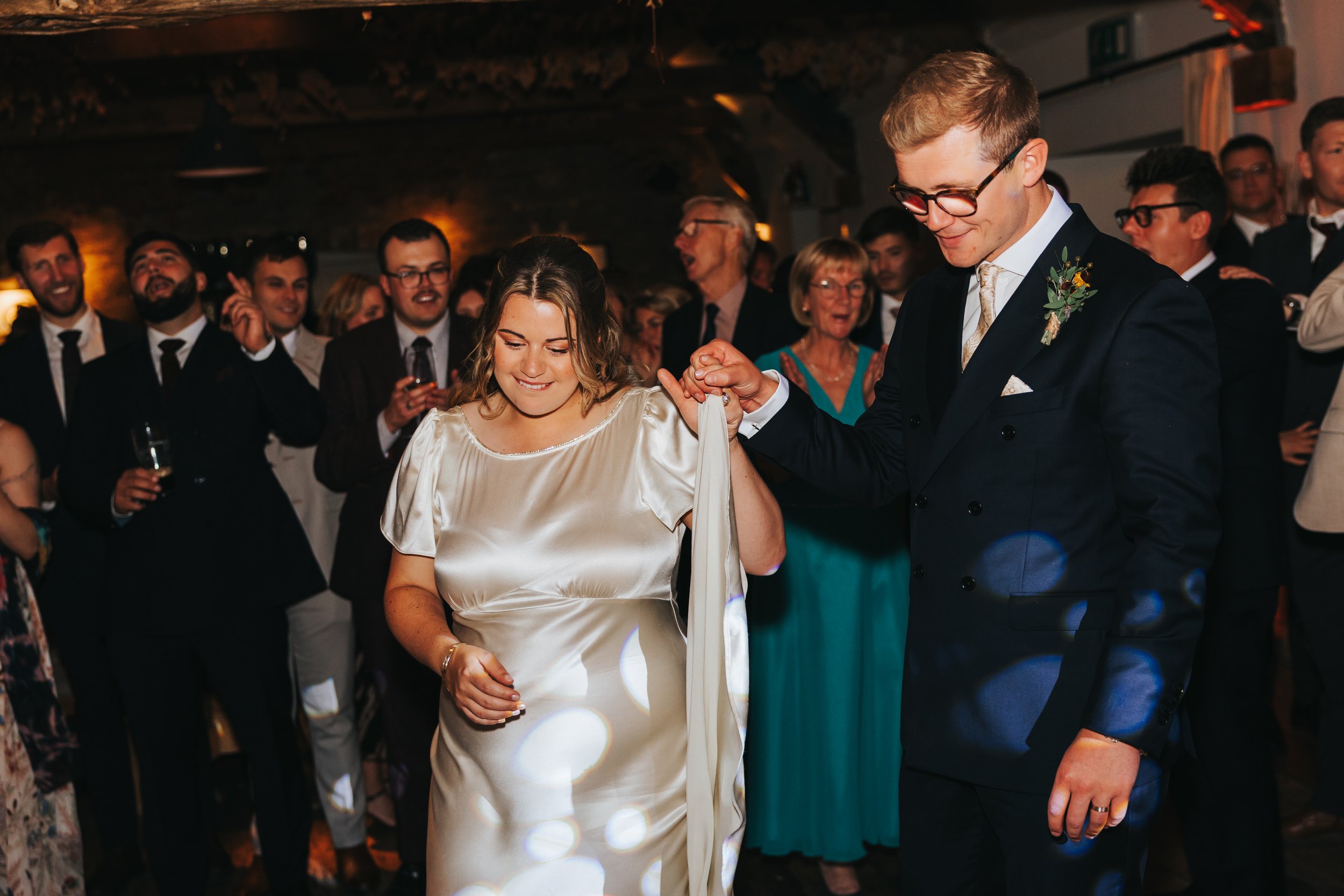 A bride and groom dance at their wedding reception, surrounded by guests in formal attire, with warm lighting and joyful expressions.