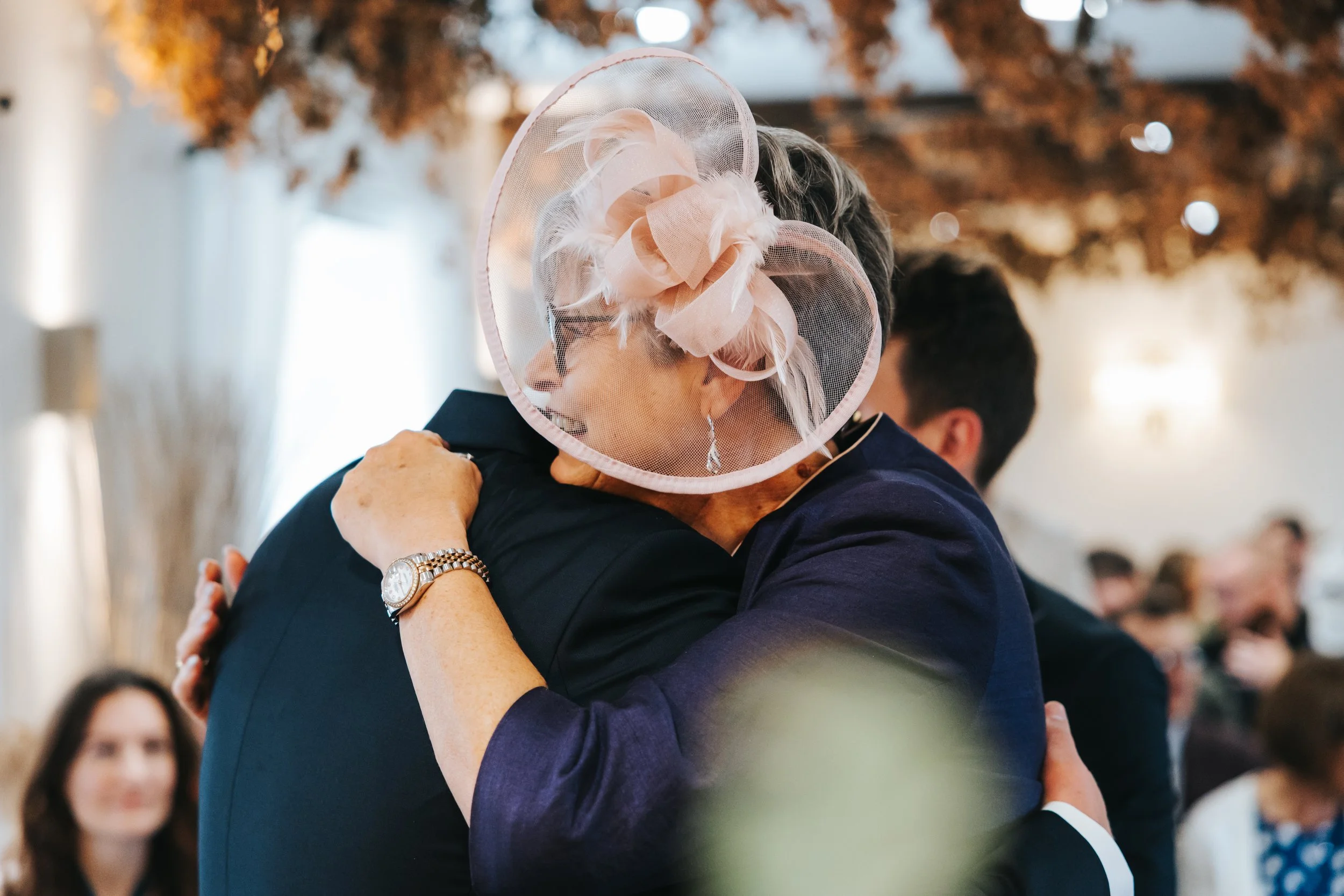 An elderly woman with a hat and earrings hugs a man in a dark suit at an indoor event, likely a wedding or celebration, with other people and decorations in the background.