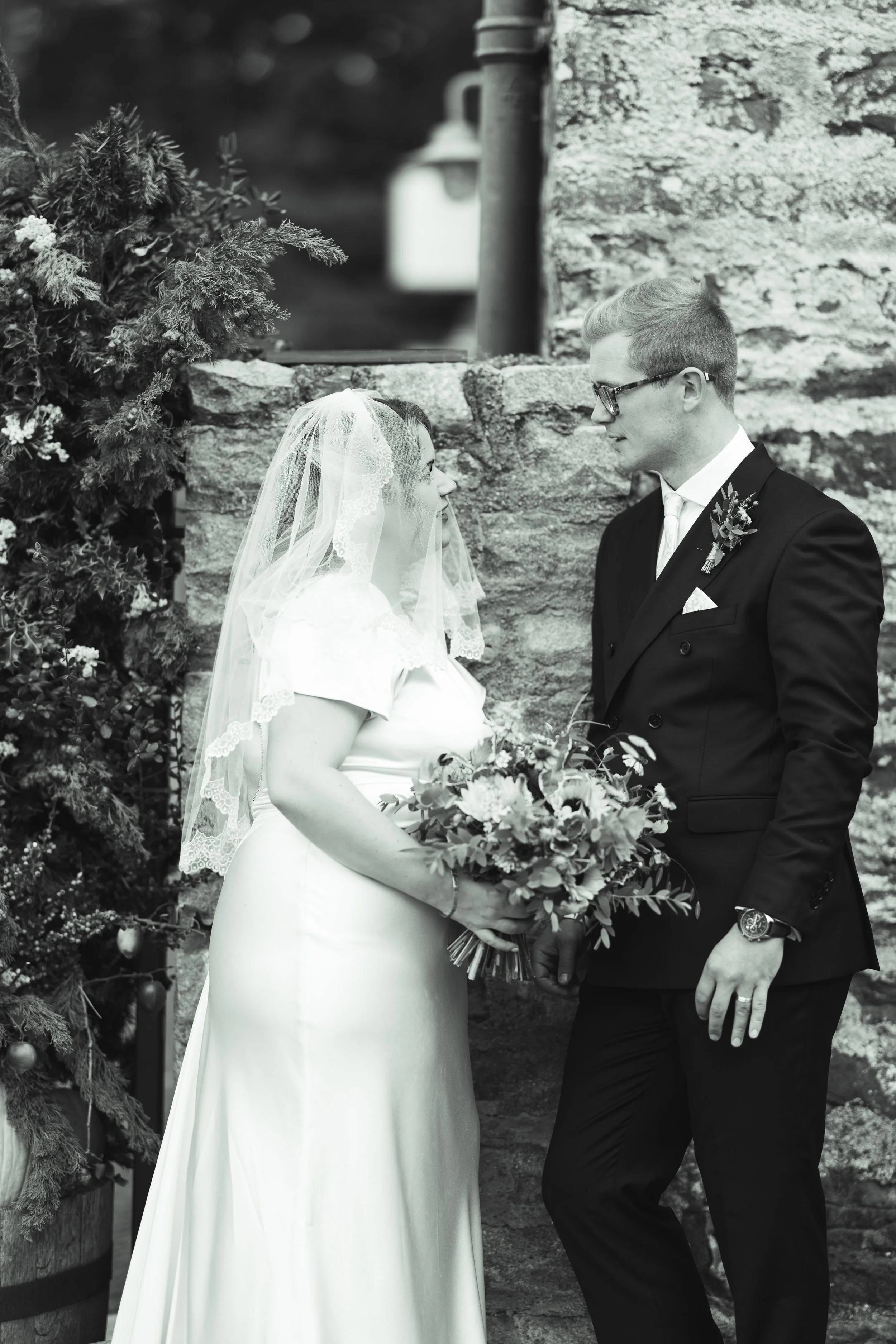 A black and white photograph of a bride and groom standing close together outdoors during their wedding. The bride is wearing a satin wedding dress with a veil, holding a bouquet of flowers, and gazing at the groom. The groom is dressed in a dark sui