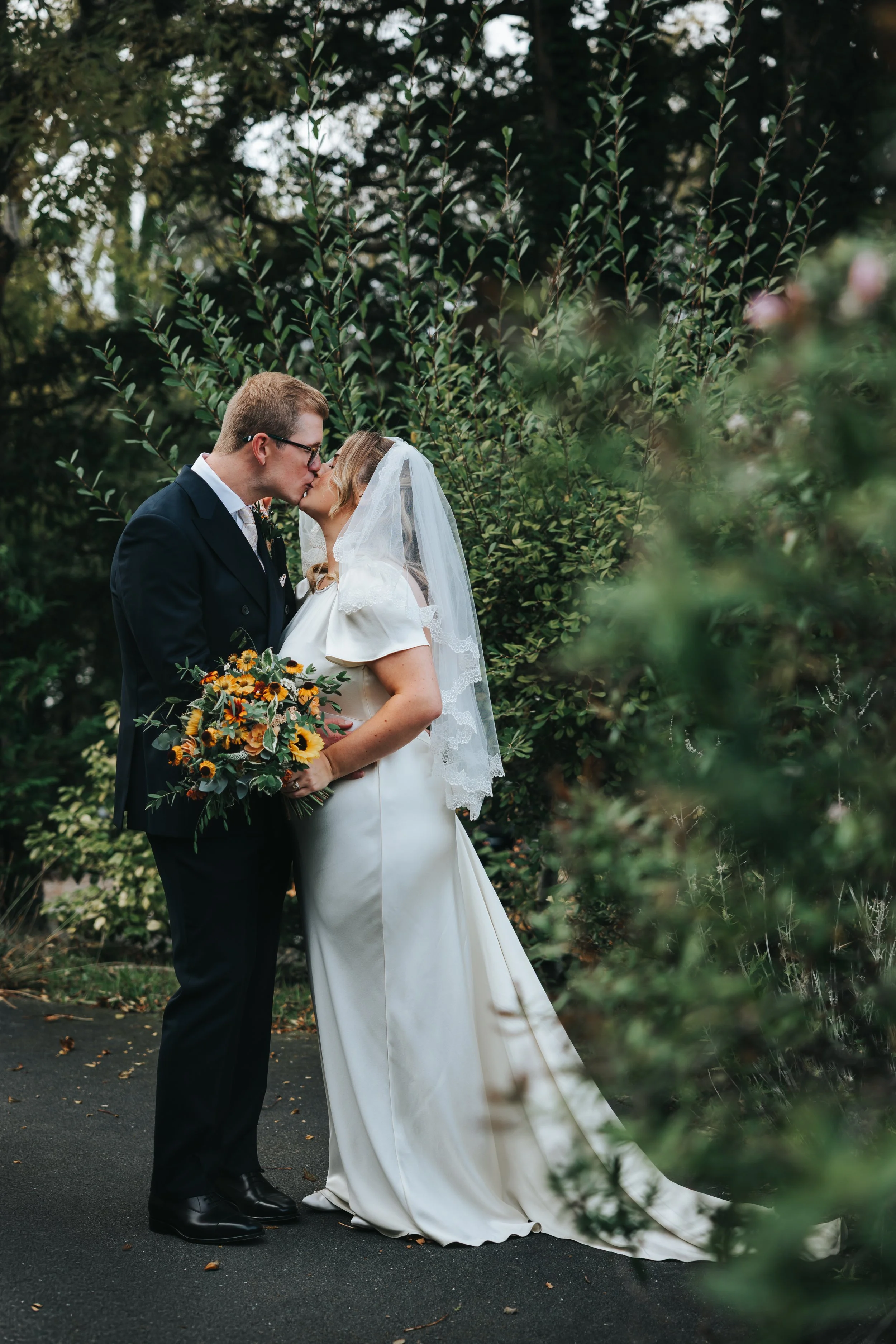 A bride and groom sharing a kiss outdoors on their wedding day, surrounded by greenery, with the bride holding a bouquet of sunflowers and other flowers.