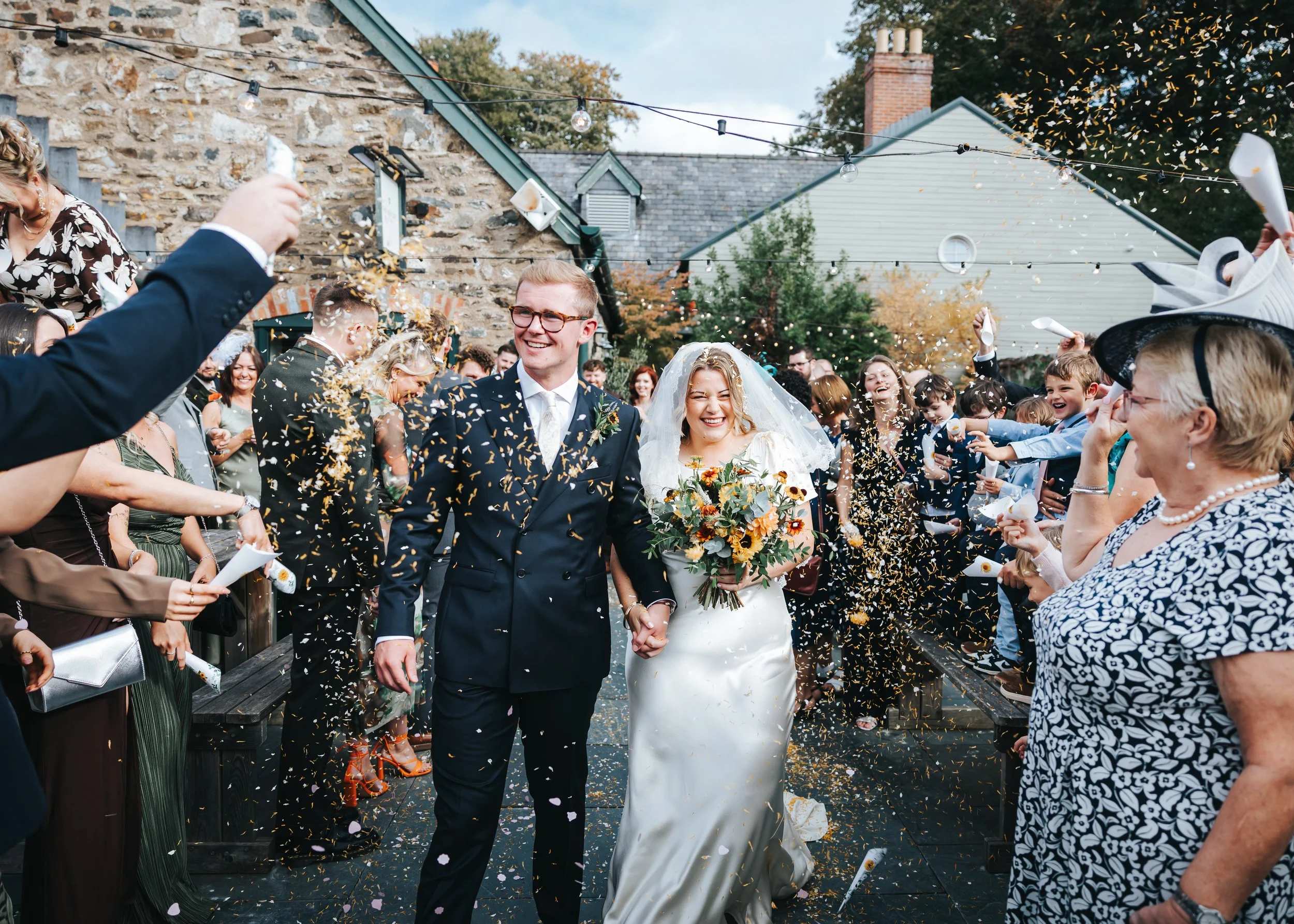 A newlywed couple walking hand in hand through a crowd of celebrating guests outdoors, with confetti in the air and warm, festive atmosphere.