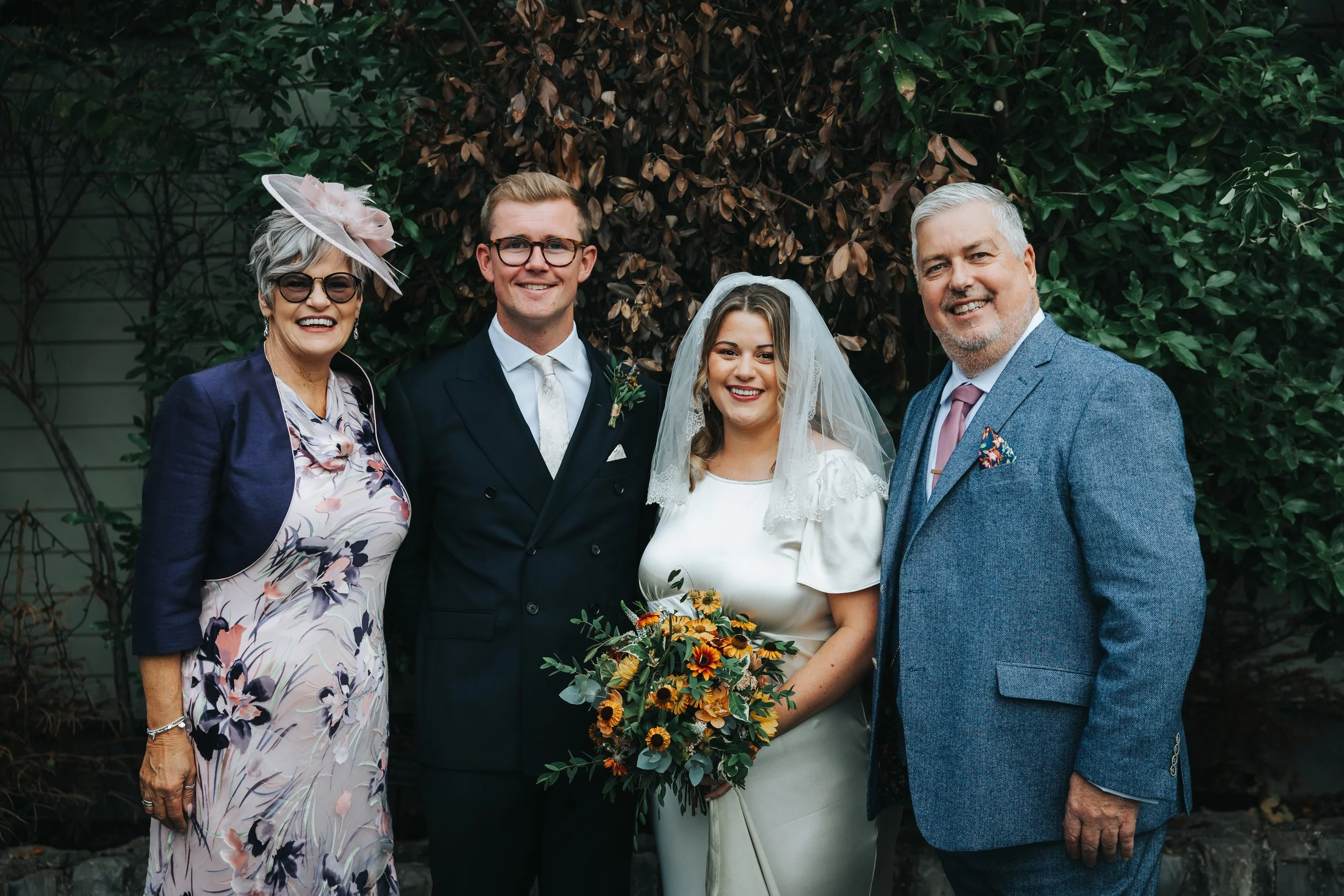 A group of five people, including a bride and groom, smiling and posing for a photo outdoors in front of greenery. The bride is holding a bouquet and wearing a white wedding dress with a veil, while the groom is in a dark suit with a white tie. Two o