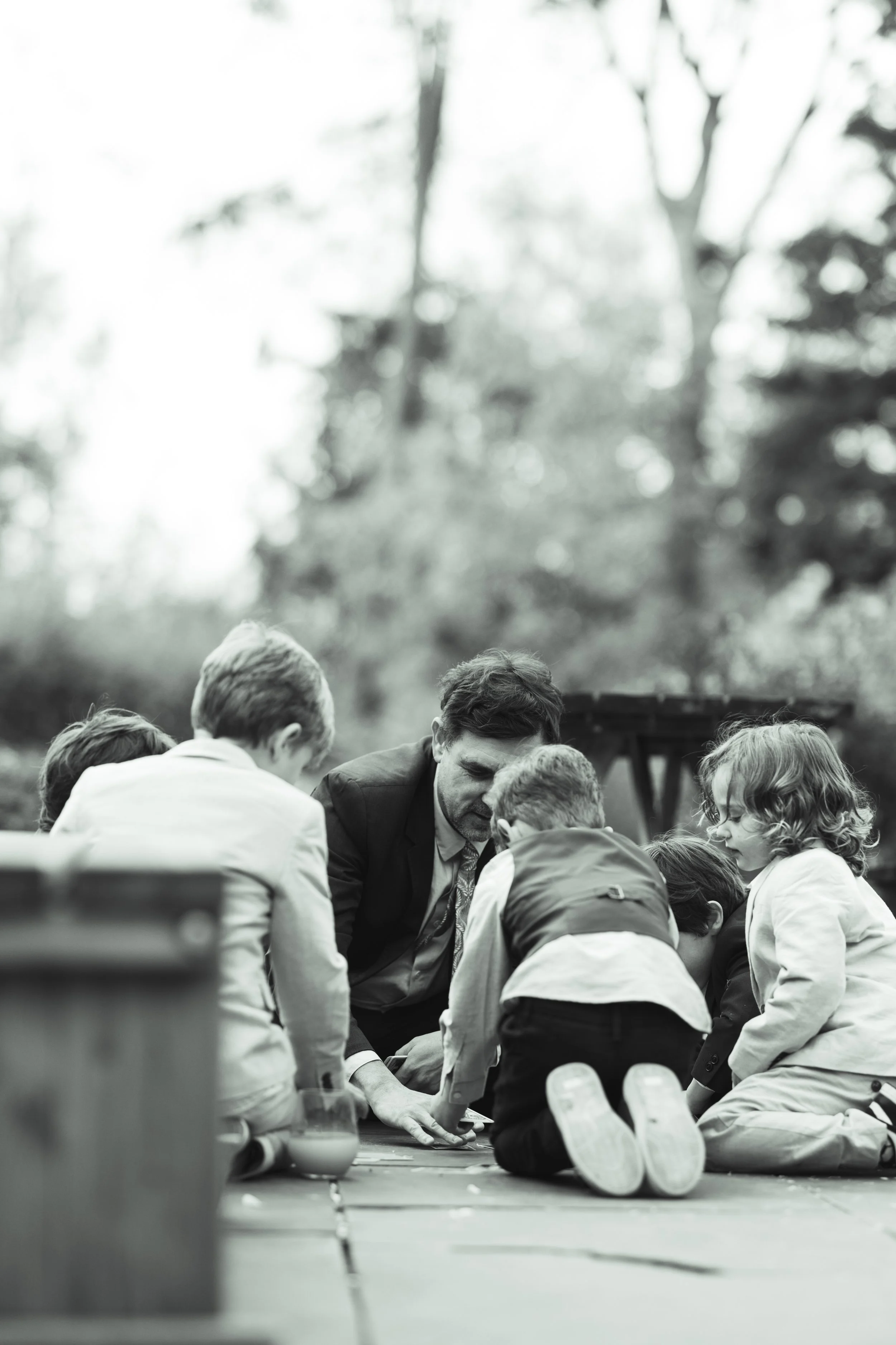 A man in a suit and five children gathered around on the ground outdoors, possibly playing a game or working on a project, with trees in the background.