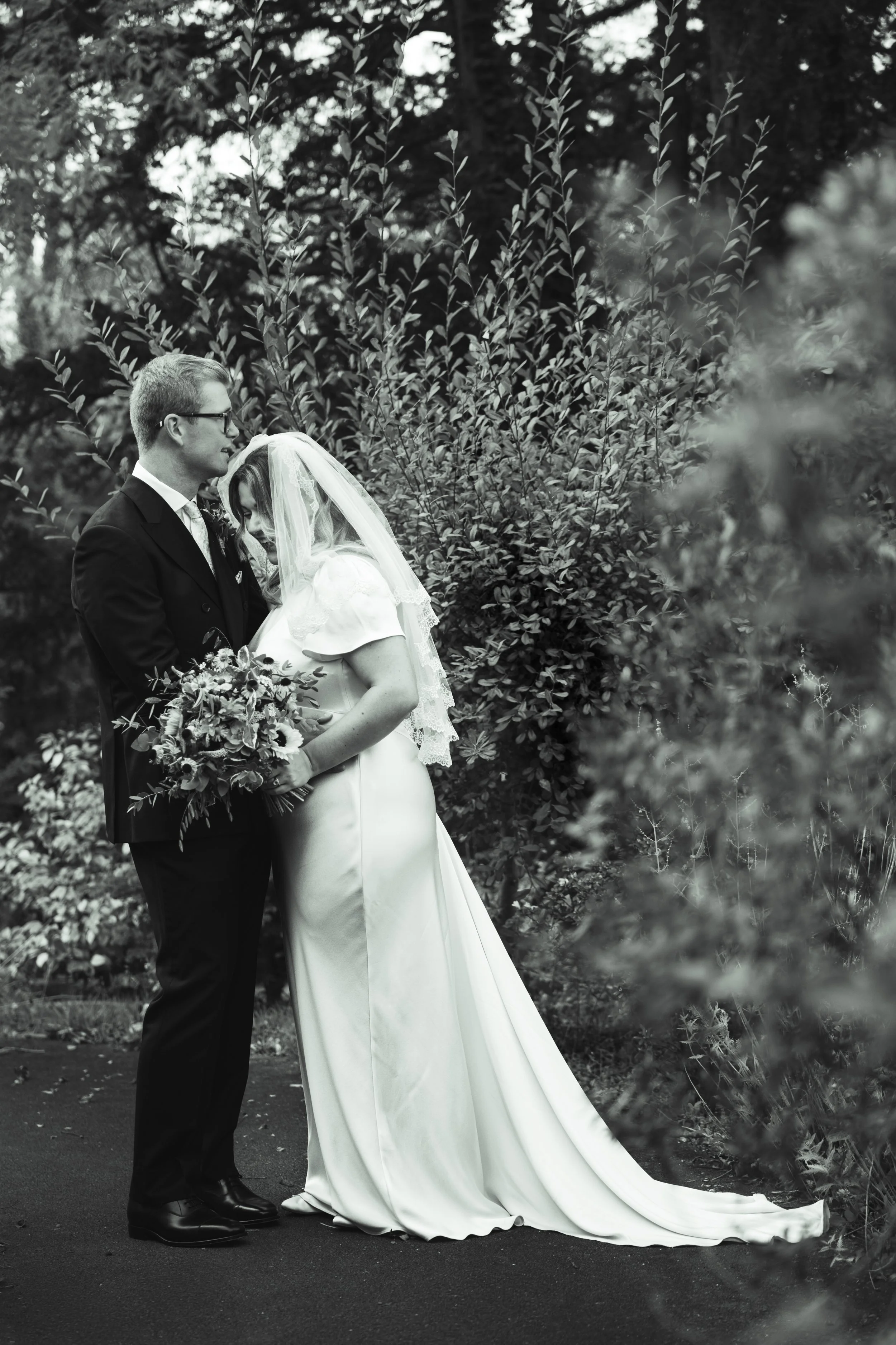 Black and white photo of a bride and groom standing close in a garden, with the groom gently resting his forehead on the bride's head. The bride is holding a bouquet and wearing a long dress with a veil. Dense foliage surrounds them.