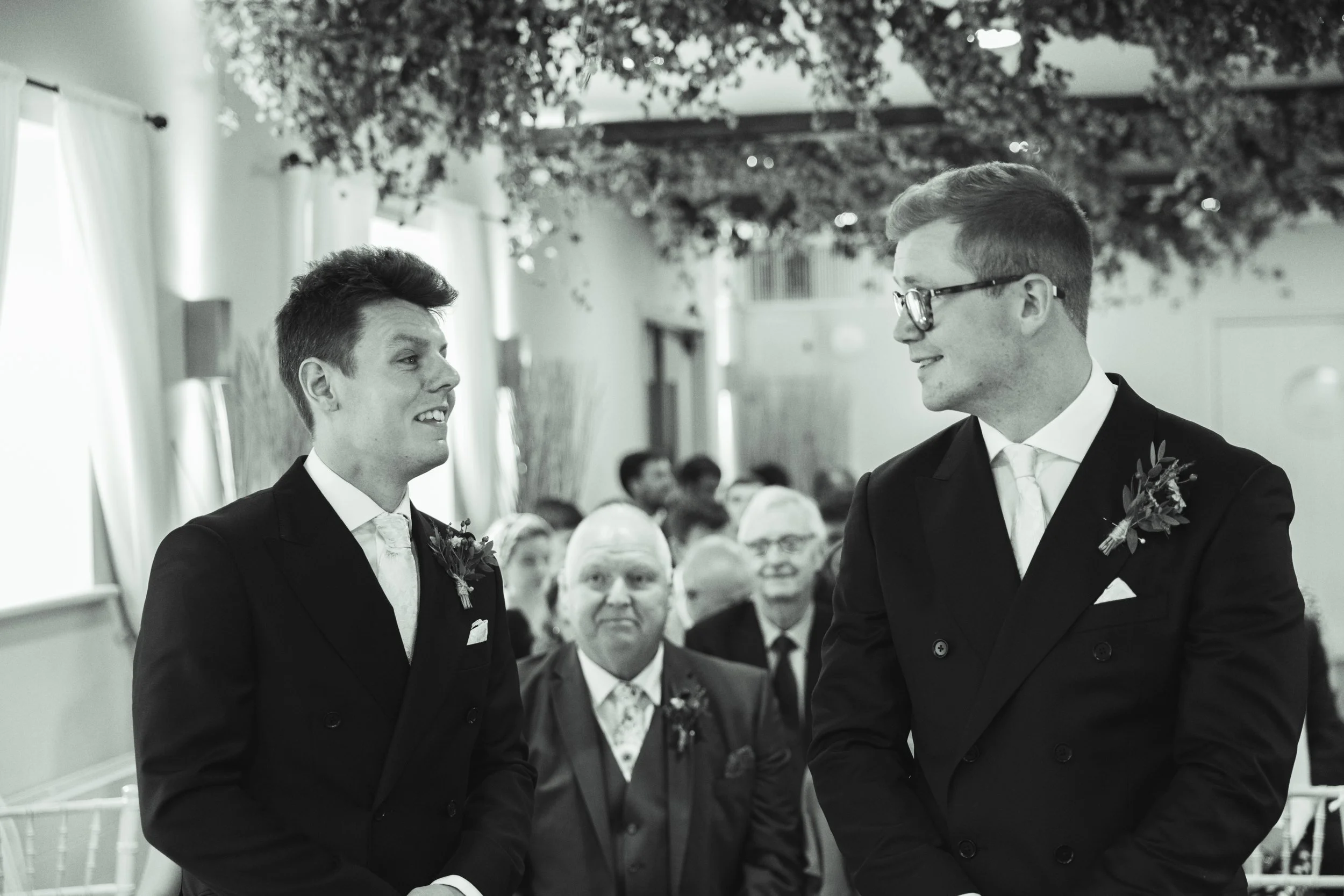 Two men in suits and glasses facing each other at a wedding ceremony, surrounded by seated guests in a decorated venue.