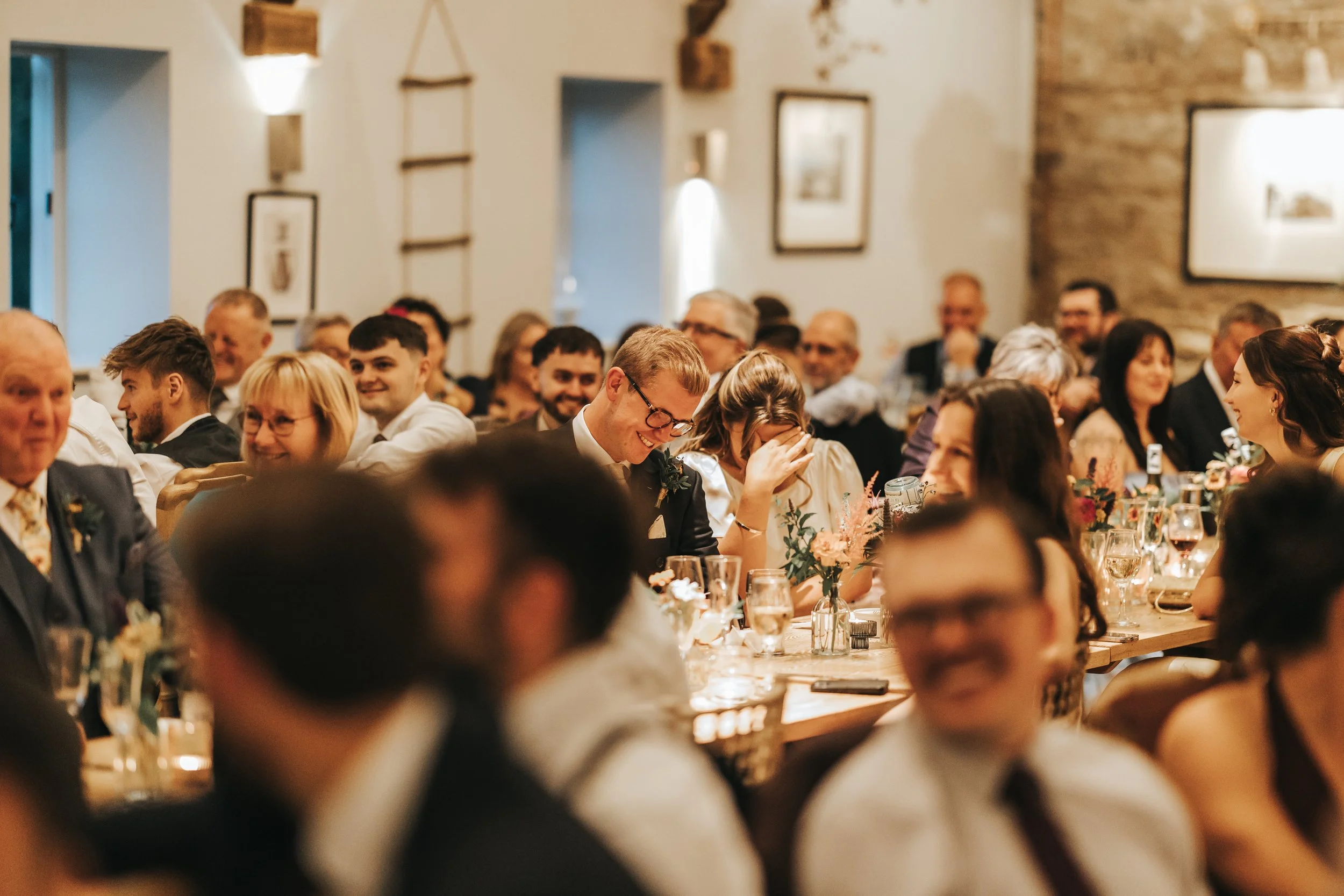 Guests at a wedding reception sitting at long tables with floral centerpieces, dressed in formal attire, smiling and laughing.