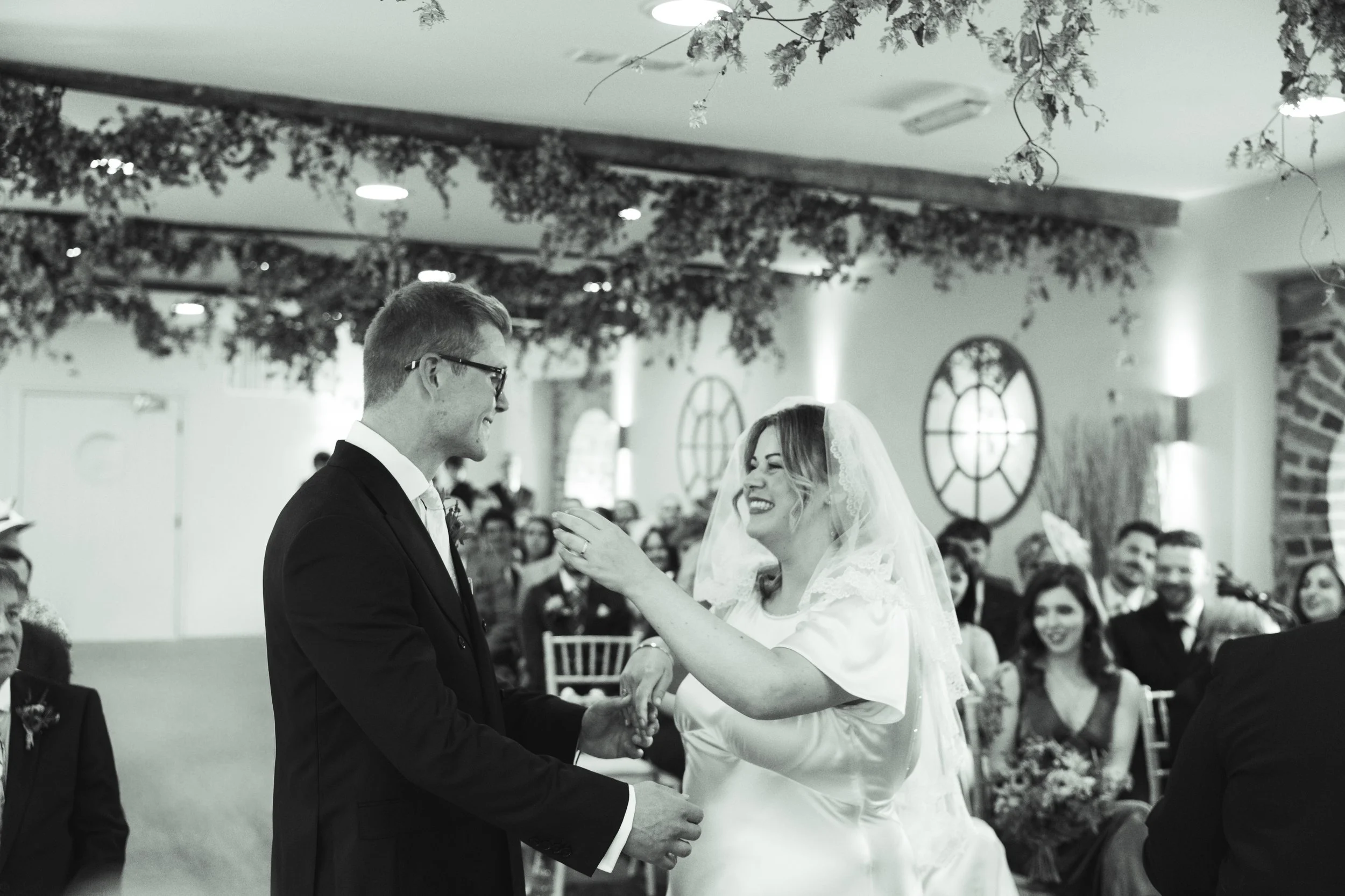 Black and white photo of a wedding ceremony with a bride and groom exchanging vows. The bride is smiling with a lace veil, and the groom wears glasses and a formal suit. Guests are seated in the background, smiling, with round windows and decorative 