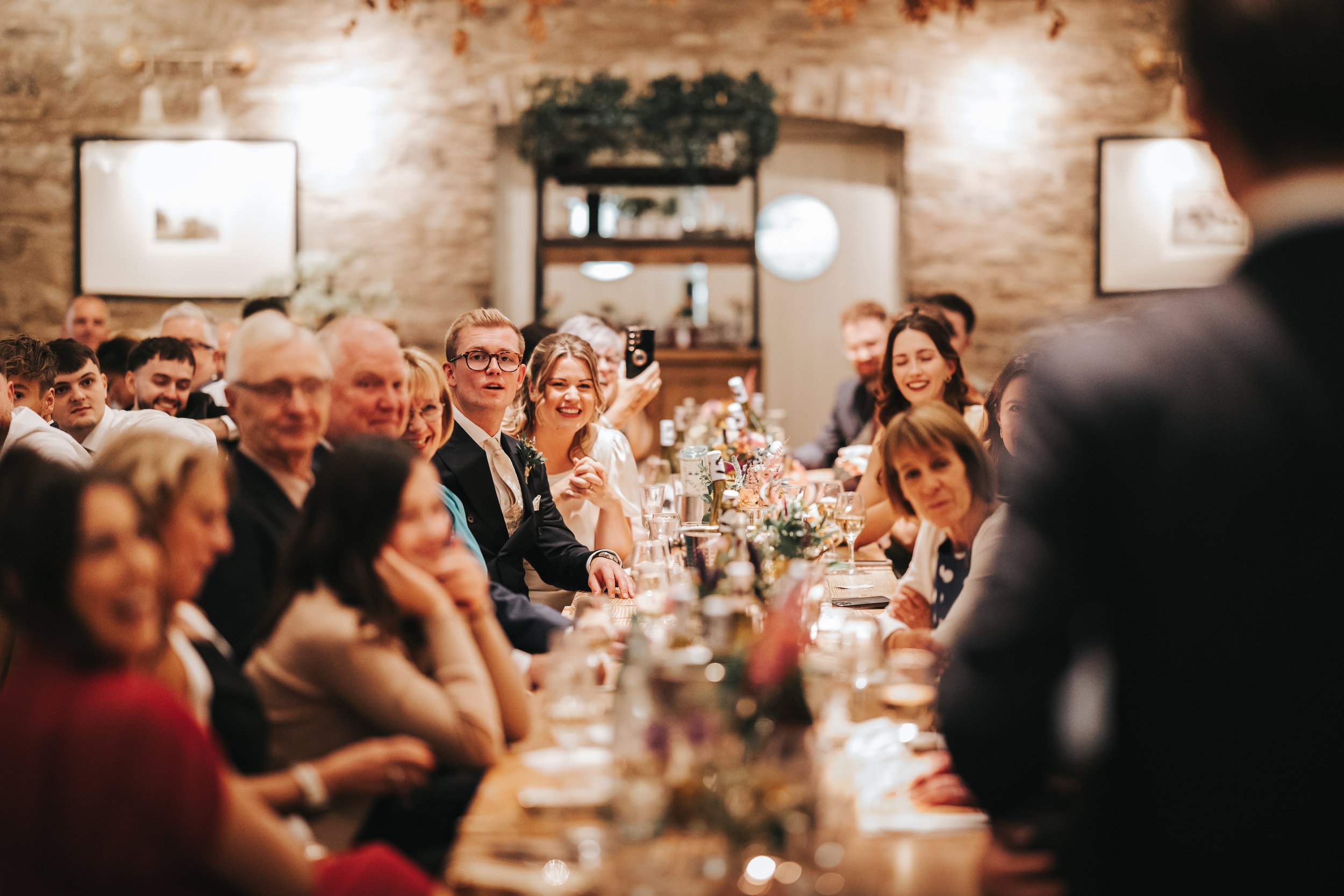 A group of people sitting at a long dinner table during a celebration or wedding reception in a cozy venue with brick walls and framed artwork, listening to a speaker or toast.