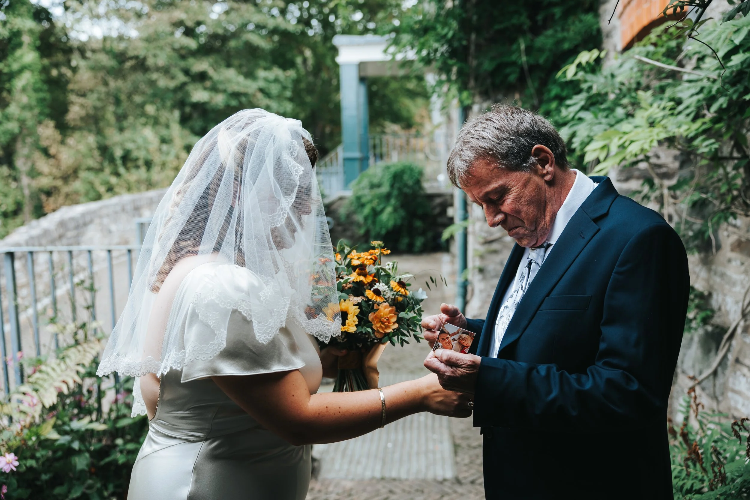 A bride in a white wedding dress and veil holding a bouquet of flowers, standing next to an older man in a dark suit, both looking at a photograph in his hand outdoors with greenery and flowers in the background.