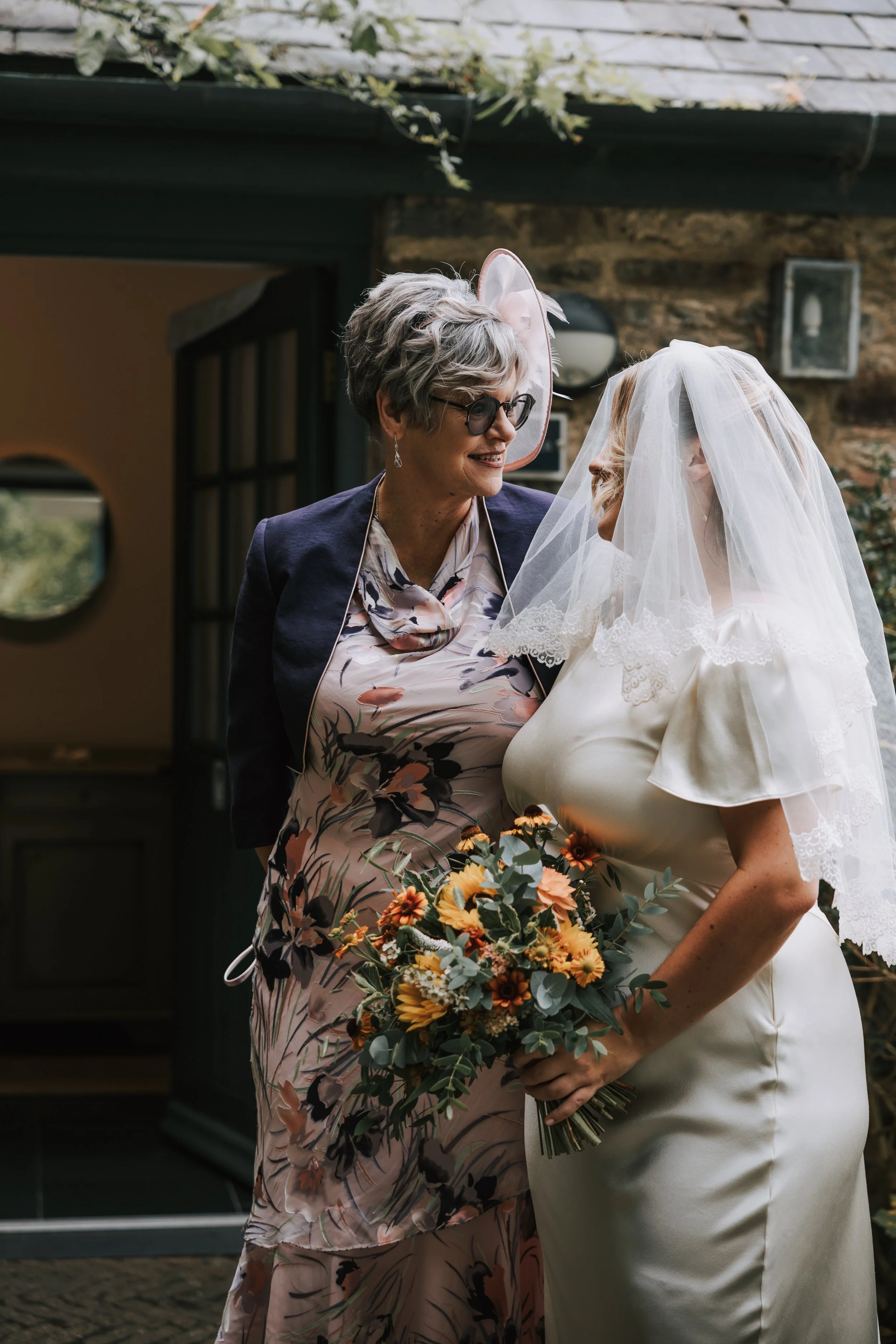 An elderly woman with gray hair and glasses, dressed in a floral dress and blazer, smiles at a bride with a veil and white dress who is holding a bouquet of yellow and orange flowers. They are outside in front of a stone building.
