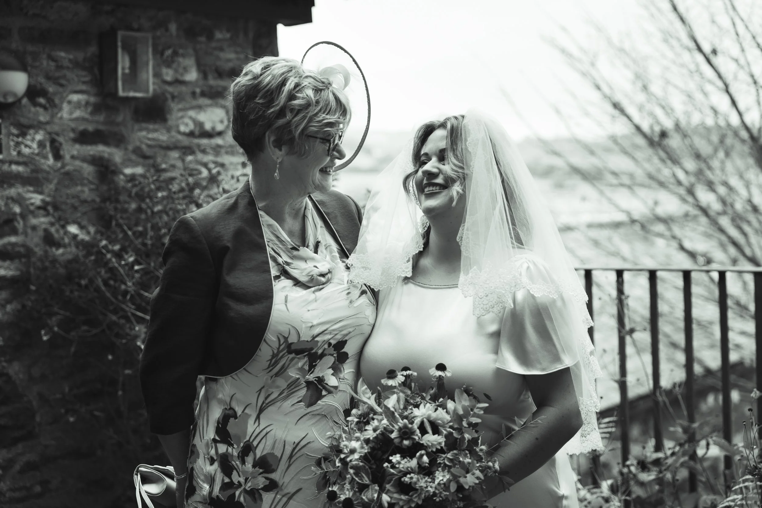 A bride and an older woman, possibly her mother, sharing a joyful moment outdoors, both smiling warmly. The bride wears a wedding dress with a lace veil and holds a bouquet, while the older woman wears glasses and a patterned blouse with a blazer.