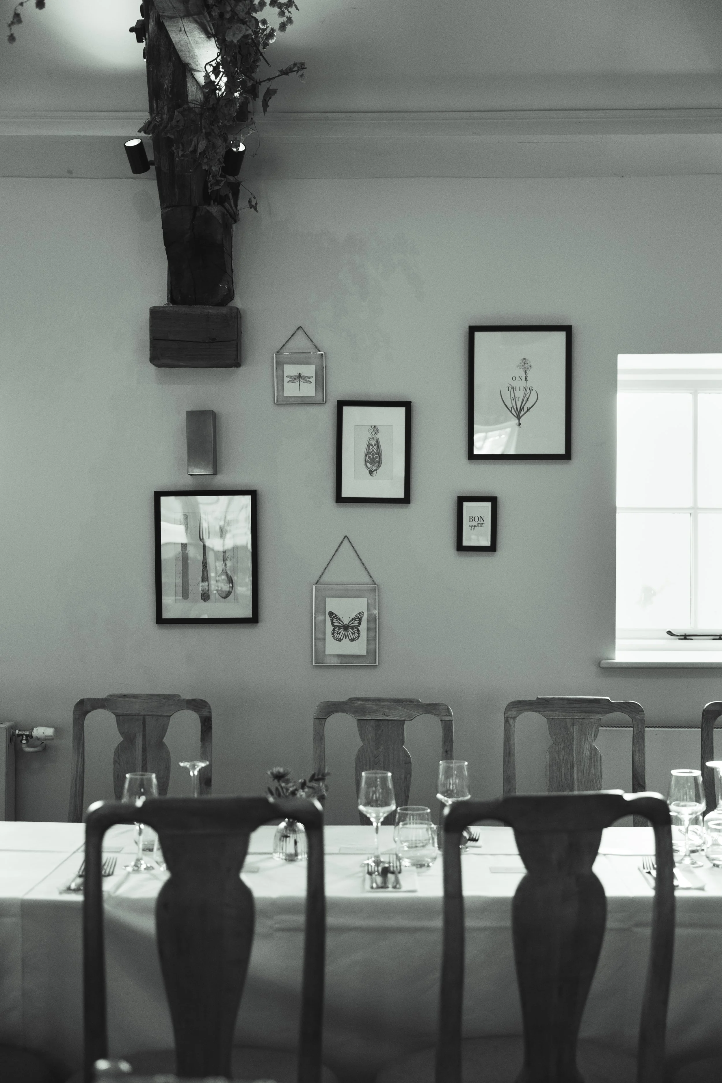 A dining room with a long table set with wine glasses, forks, and plates, surrounded by wooden chairs. The wall above has a collection of framed art and photographs, including a butterfly, a dragonfly, and various abstract designs. There's a window o