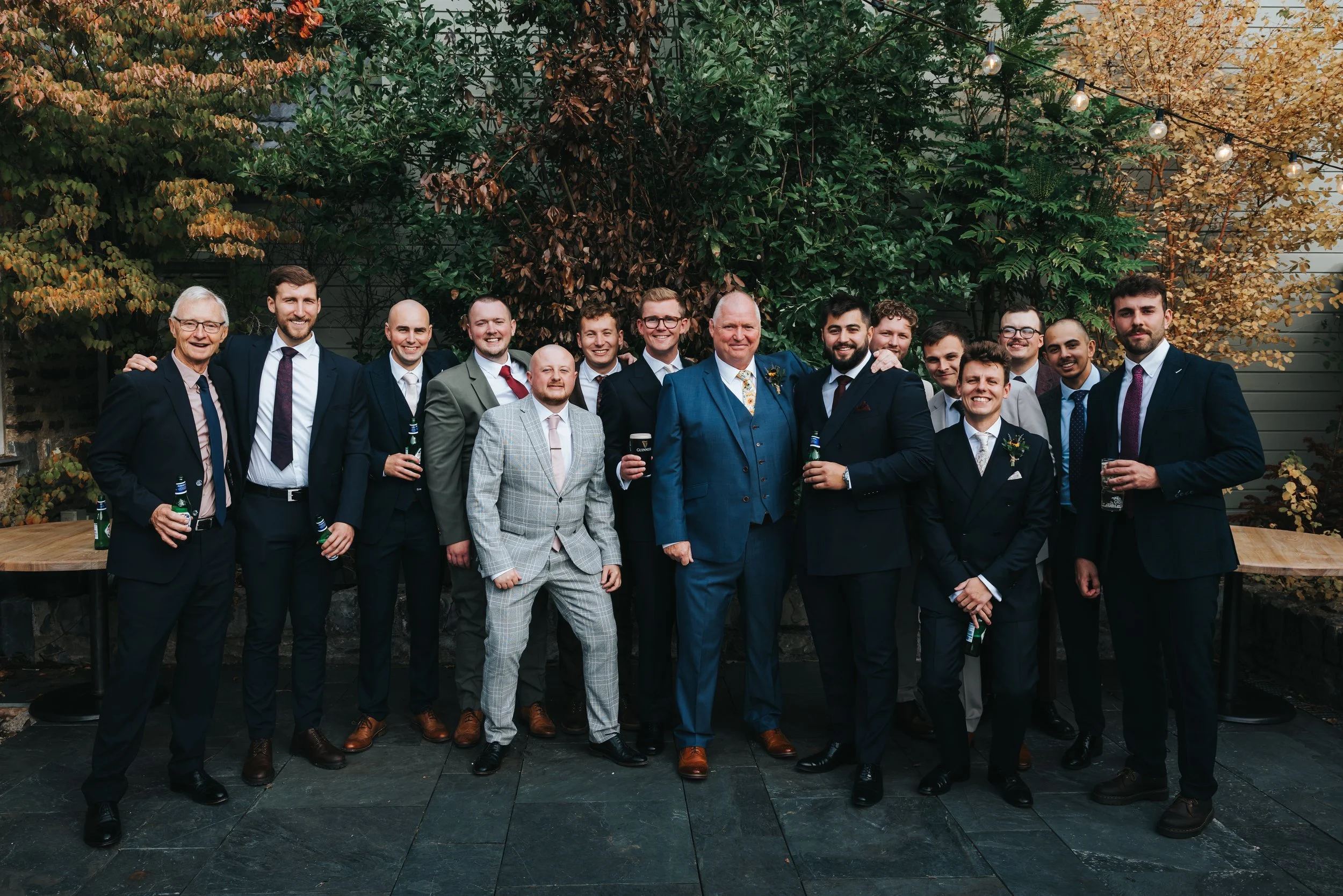 Group of men dressed in suits, celebrating outdoors during autumn, with colorful foliage and string lights overhead.