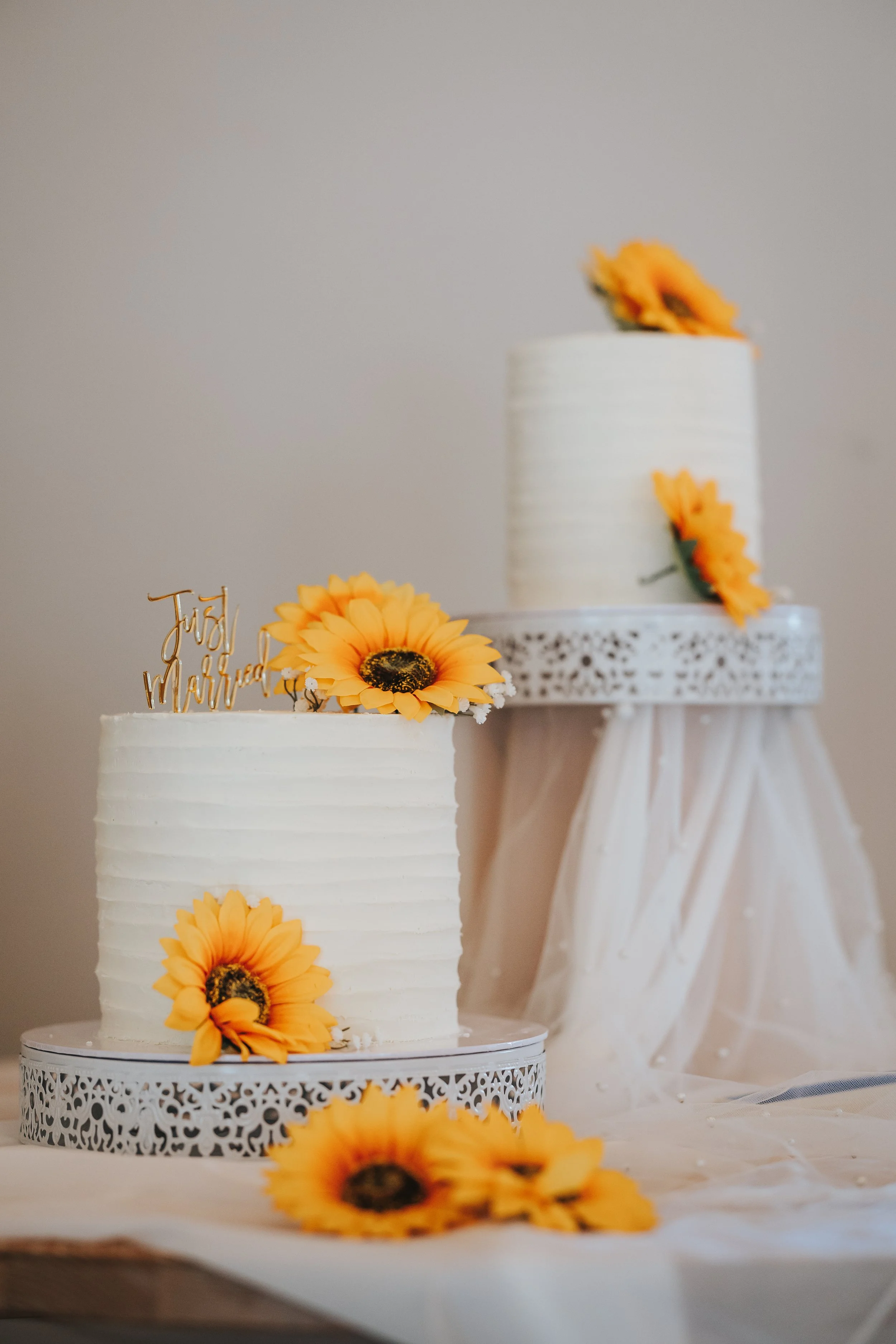 Two tiered white wedding cakes decorated with yellow sunflowers and a gold "Just Married" topper, placed on ornate white cake stands on a draped table.