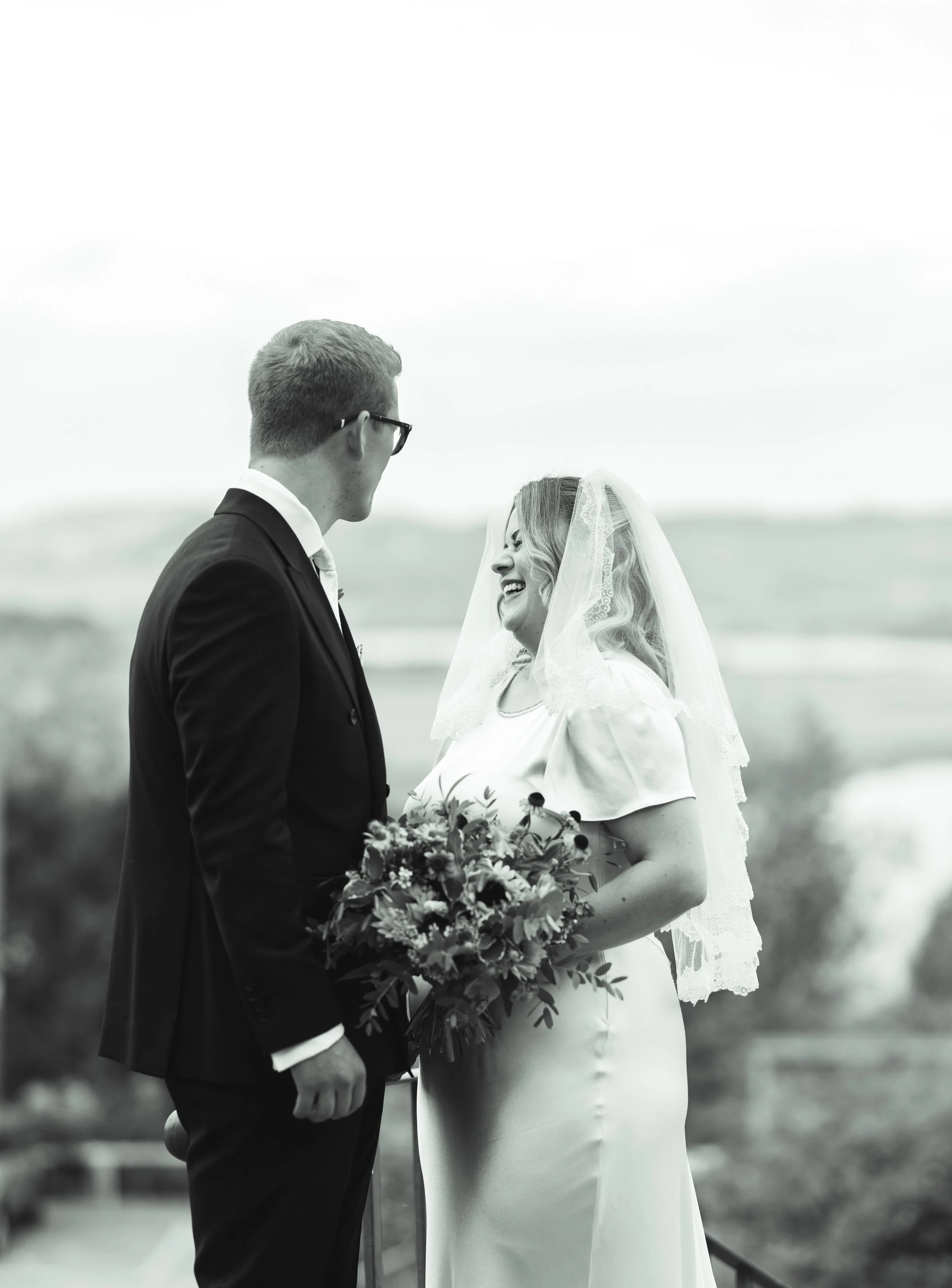 Black and white photo of a bride and groom sharing a moment outdoors. The bride is smiling, holding a bouquet, and wearing a lace veil, while the groom is looking at her. The background shows a scenic landscape.