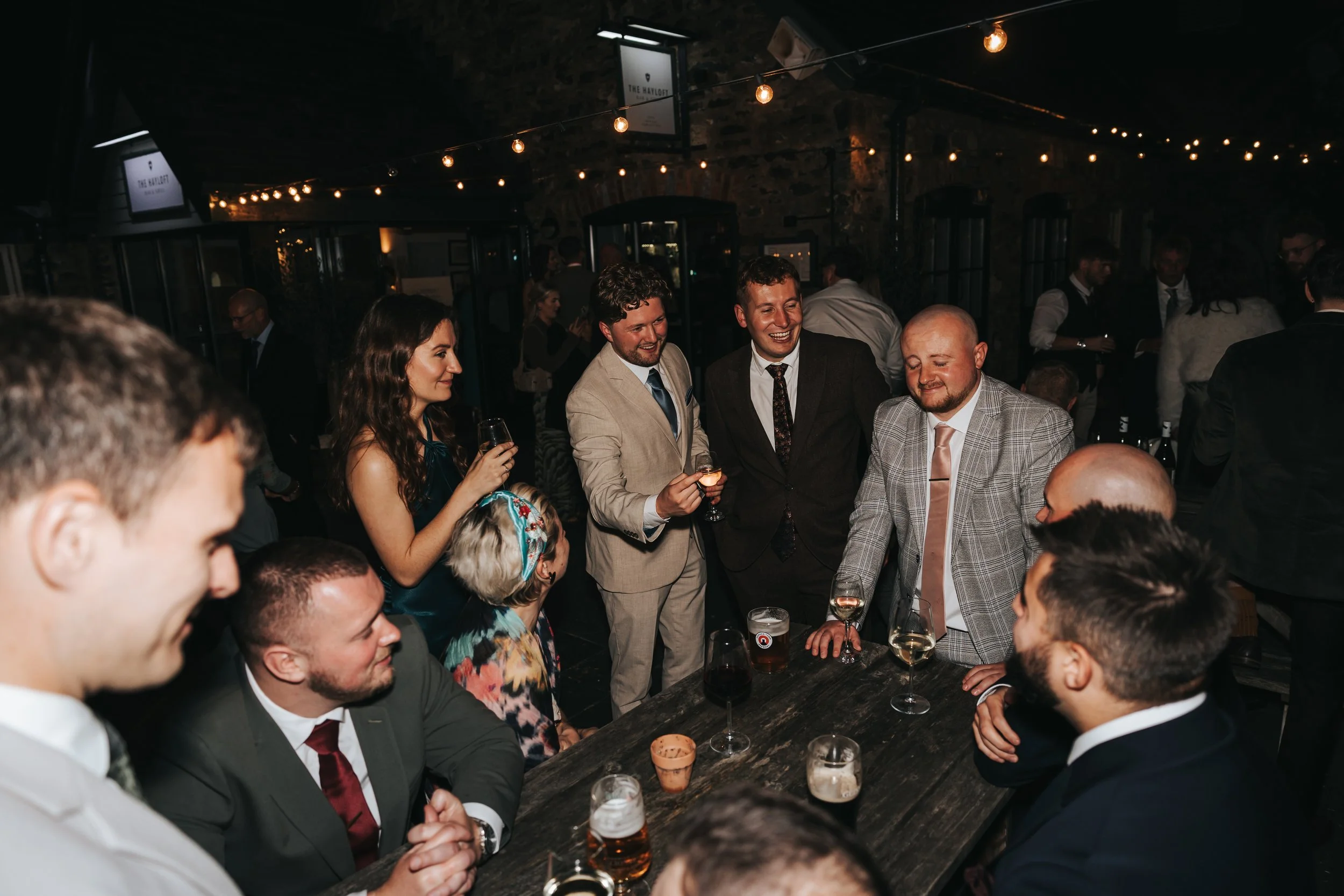 Group of people at a social gathering in a dimly lit venue, wearing formal attire, with drinks on the table and engaging in conversation and laughter.