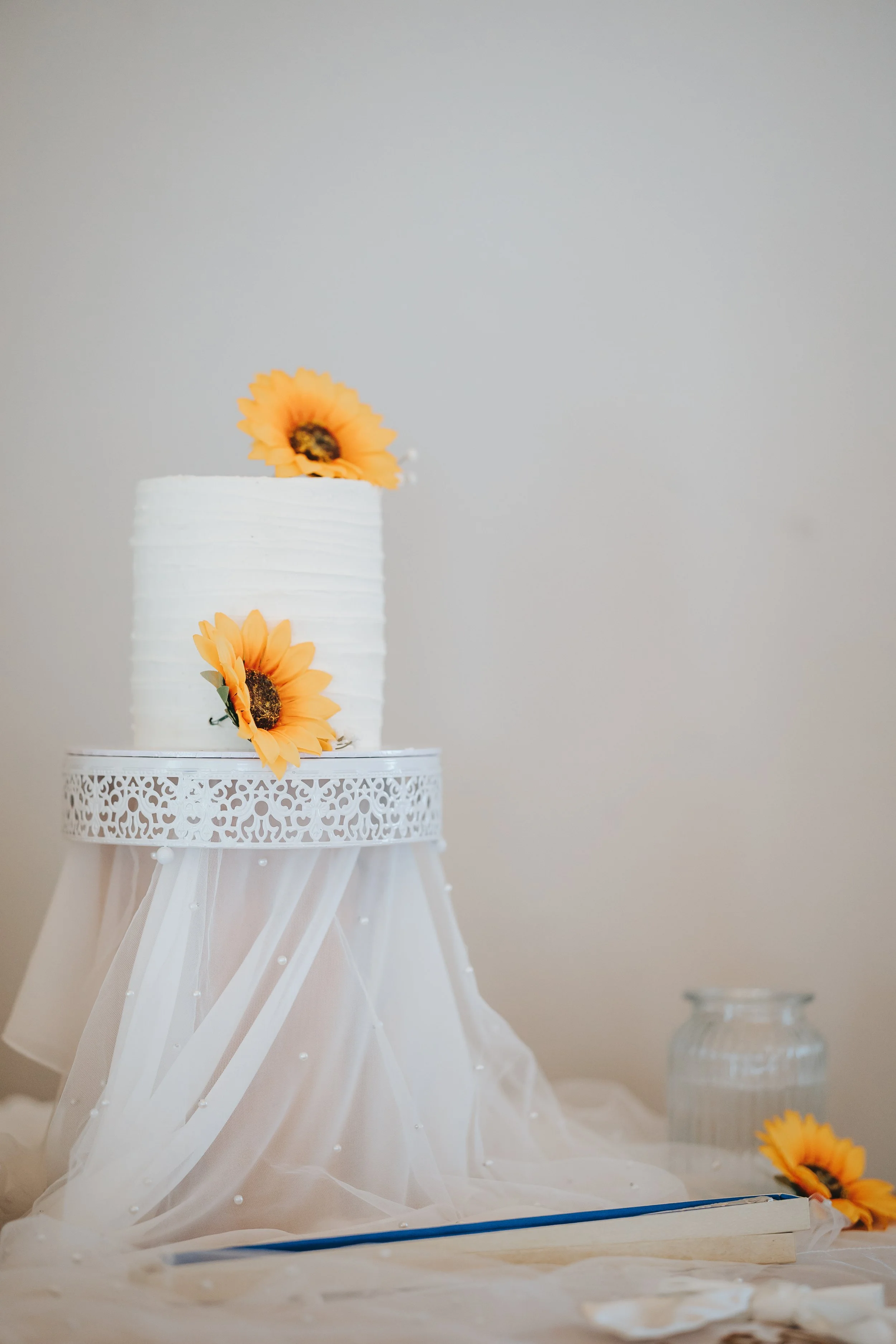 A white wedding cake decorated with sunflower flowers, placed on a decorative table draped with white fabric and pearls, with a glass jar and sunflower on the table.