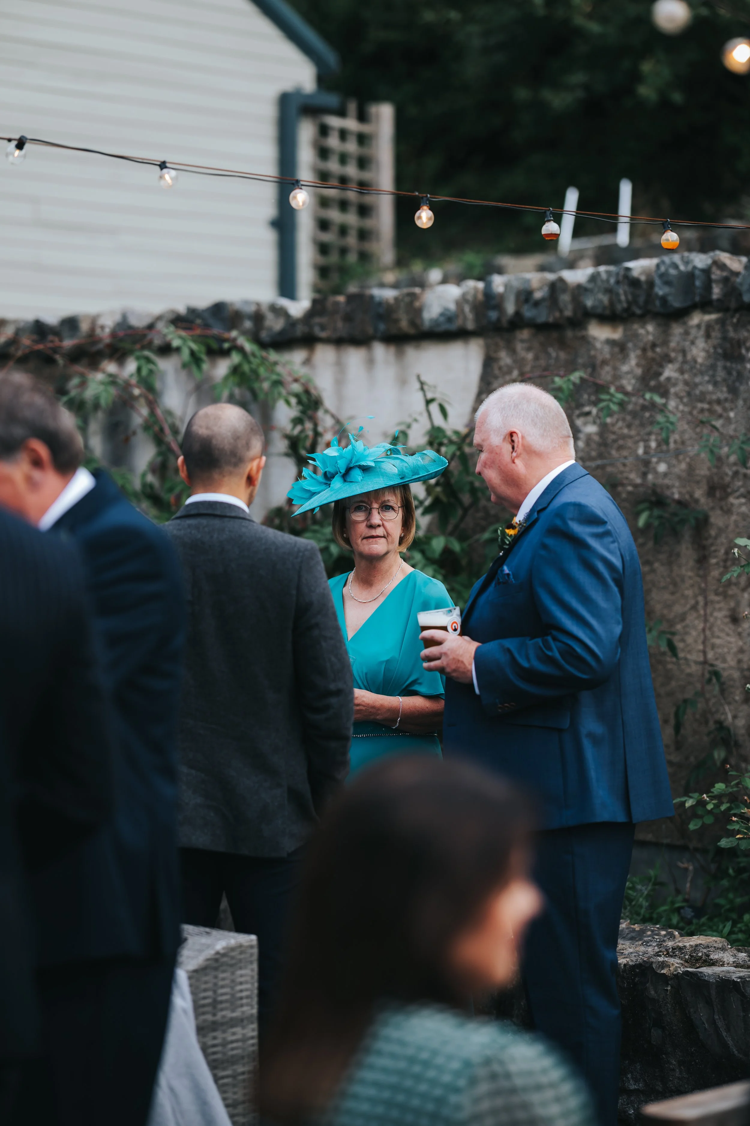 A group of people at an outdoor social event, with a woman wearing a large teal hat and dress, and men in suits, standing near a stone wall with string lights overhead.